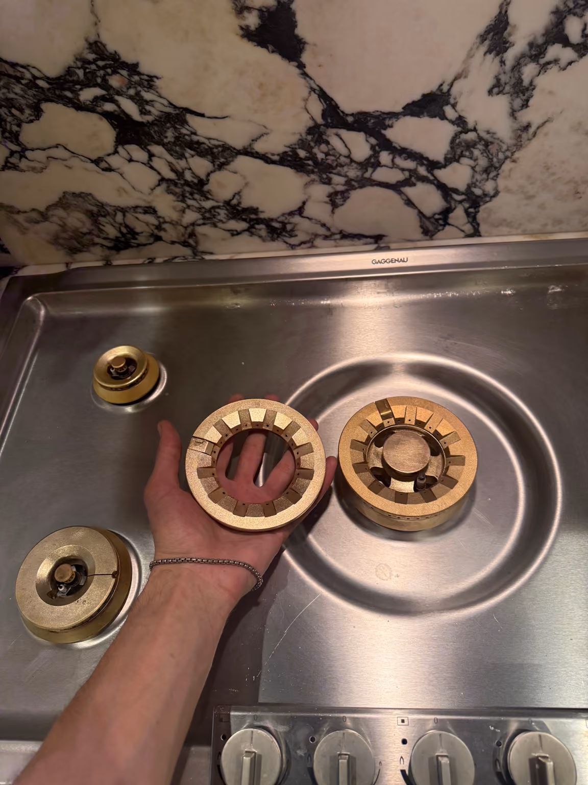 Hand holding a brass burner ring above a stainless steel cooktop.