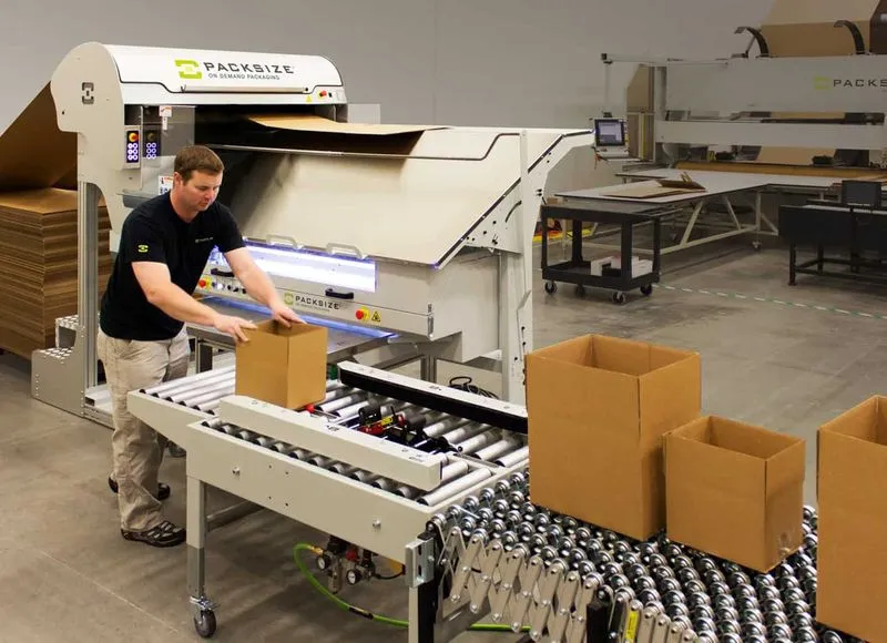 Ein Bild eines Mannes, der Kartons nach Maß vor einer Packsize iQ Verpackungsmaschine verpackt.  
