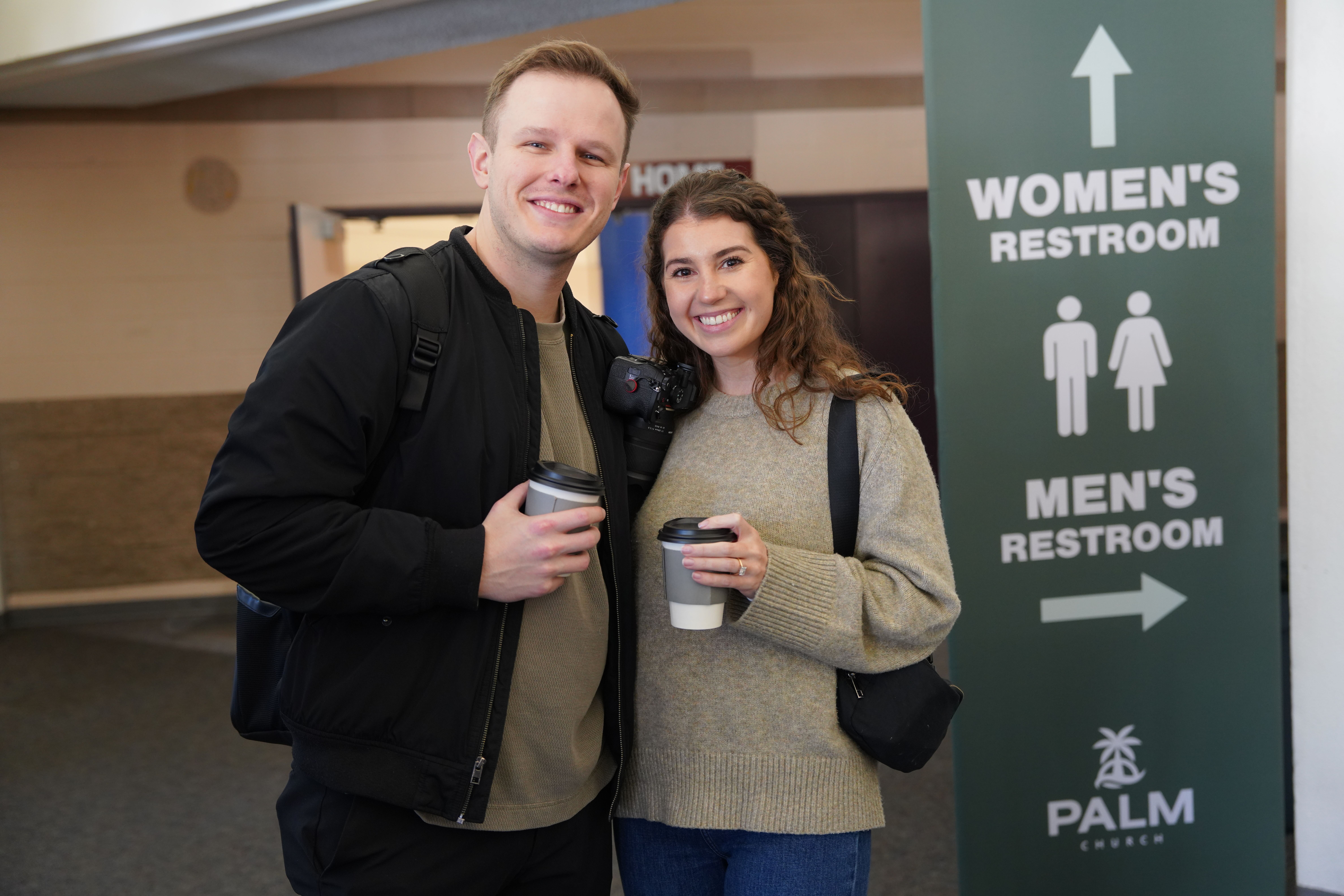 Smiling man and woman holding coffee cups standing indoors near a sign directing to women's and men's restrooms at Palm Church.