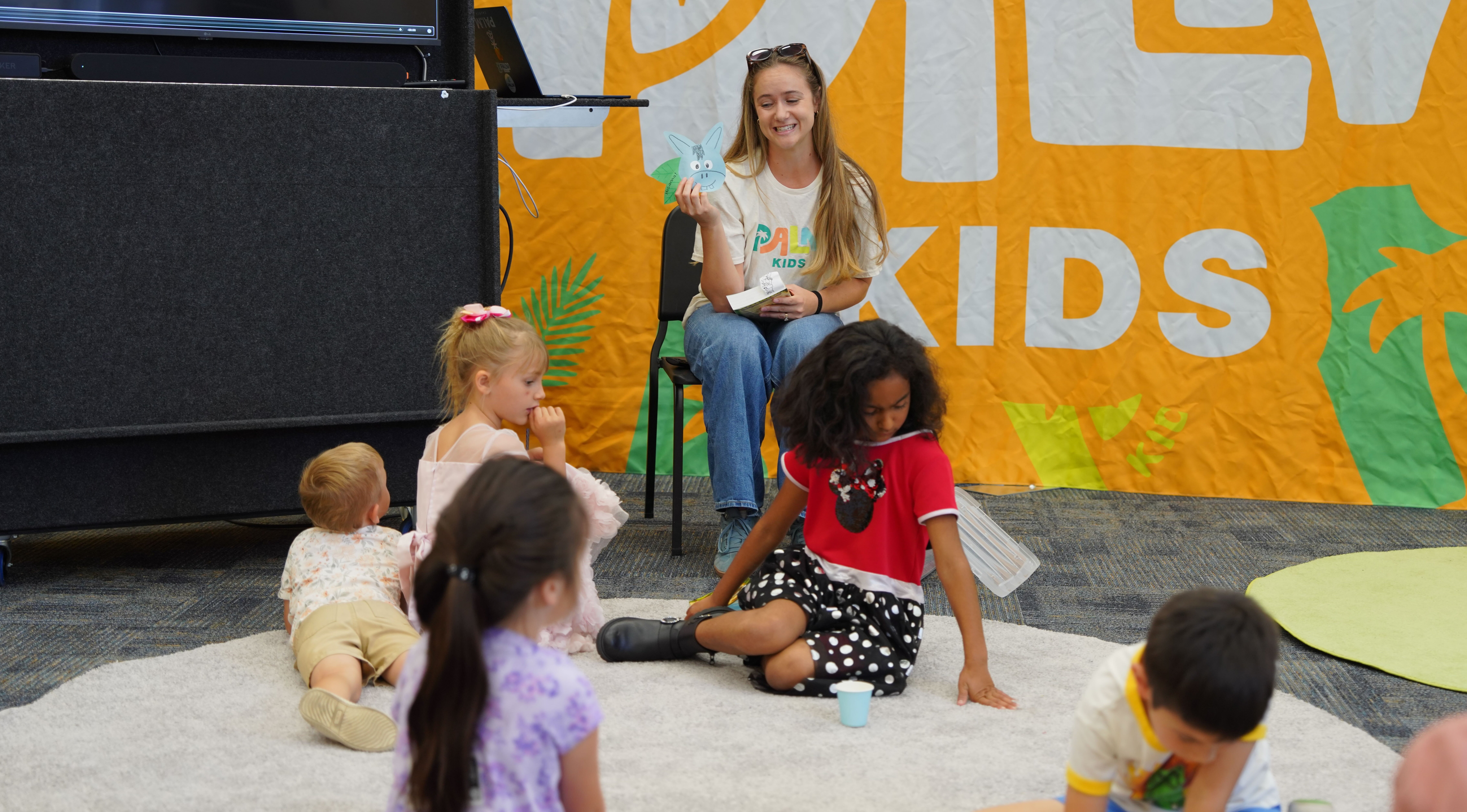 A woman reads a book and shows a paper bunny puppet to a group of young children sitting and lying on a carpeted floor during a kids' storytime session.