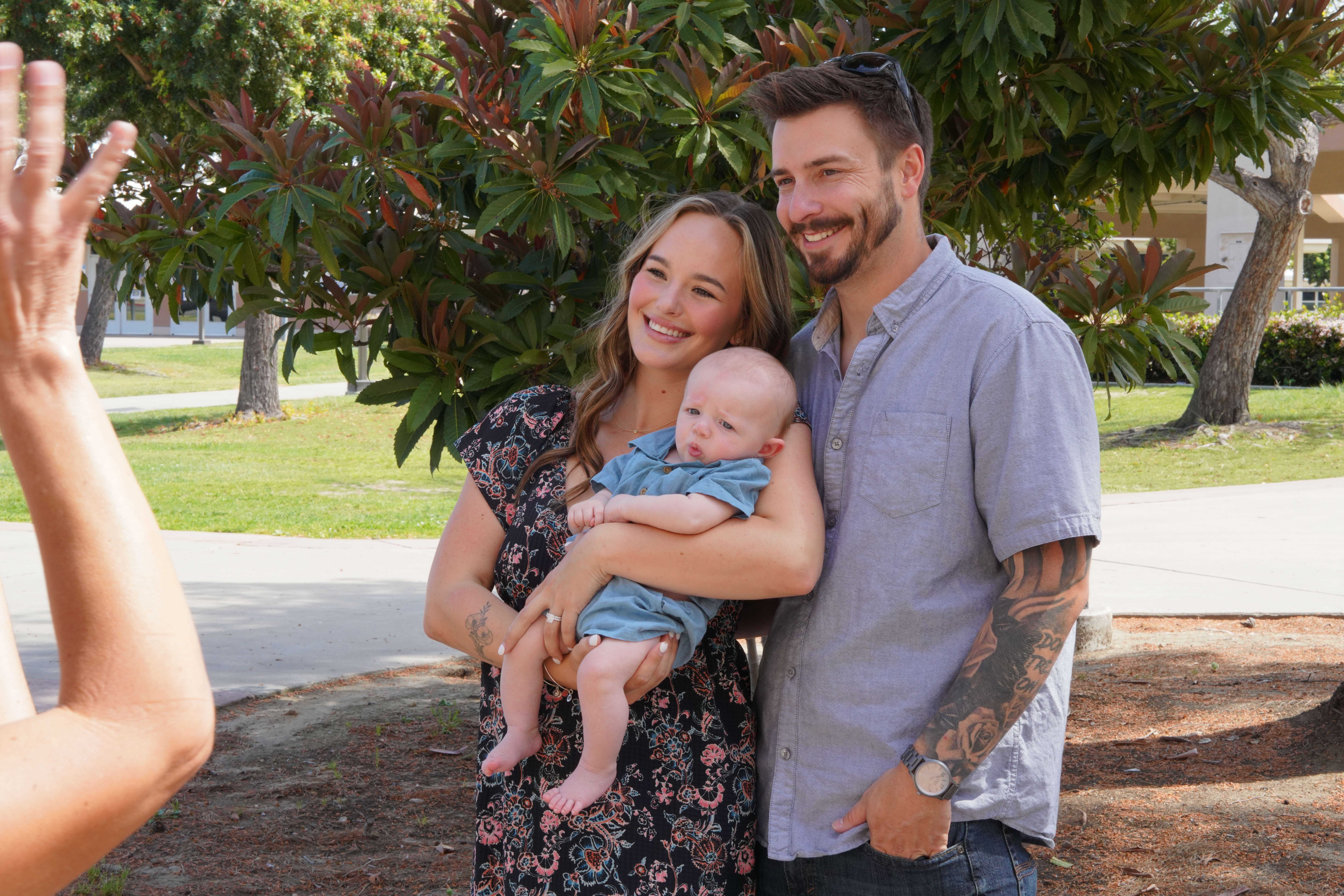 Smiling couple outdoors holding a baby while someone takes their photo.