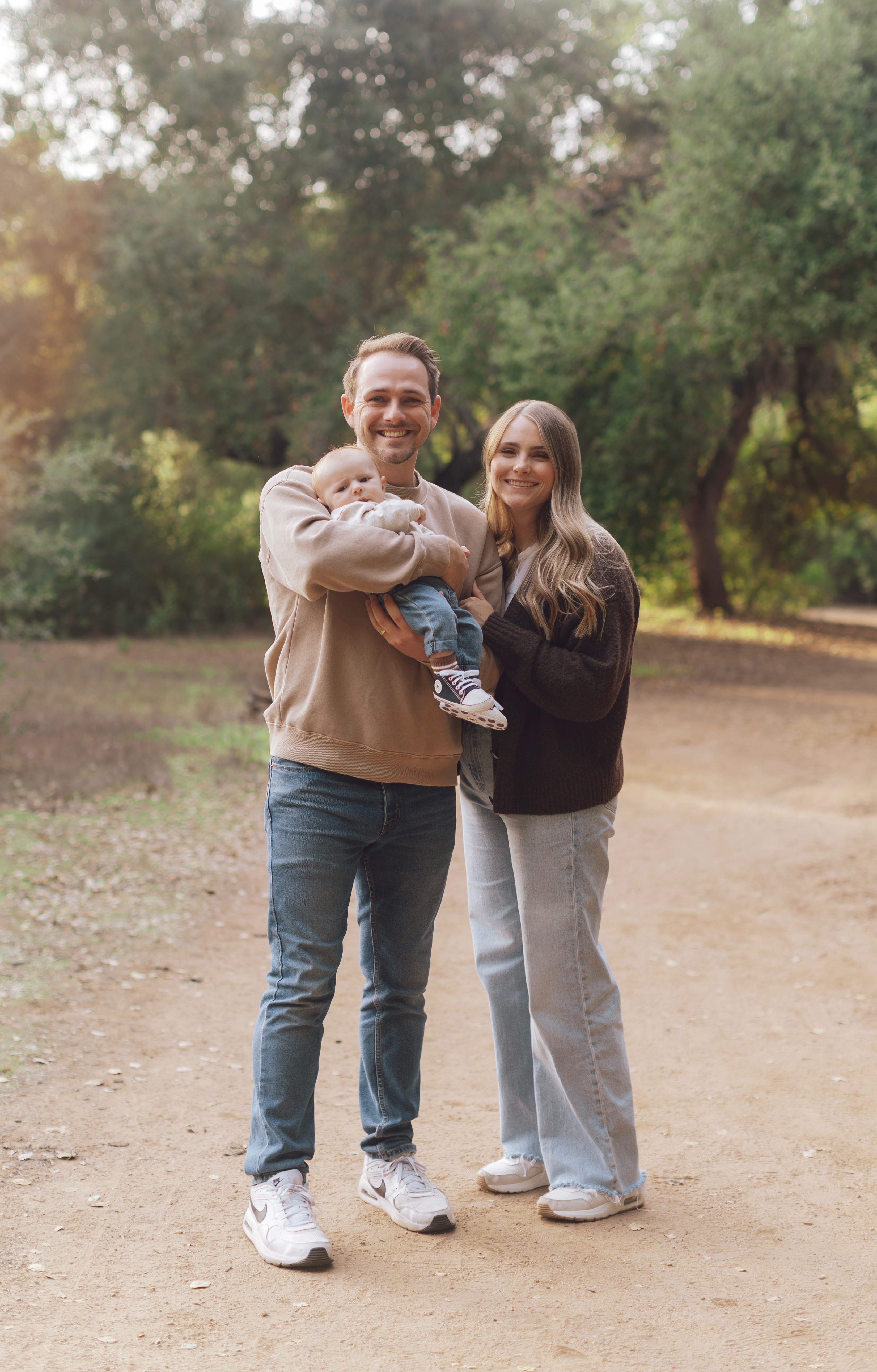 Smiling young couple standing on a dirt path in a park, with the man holding a baby wearing jeans and sneakers.