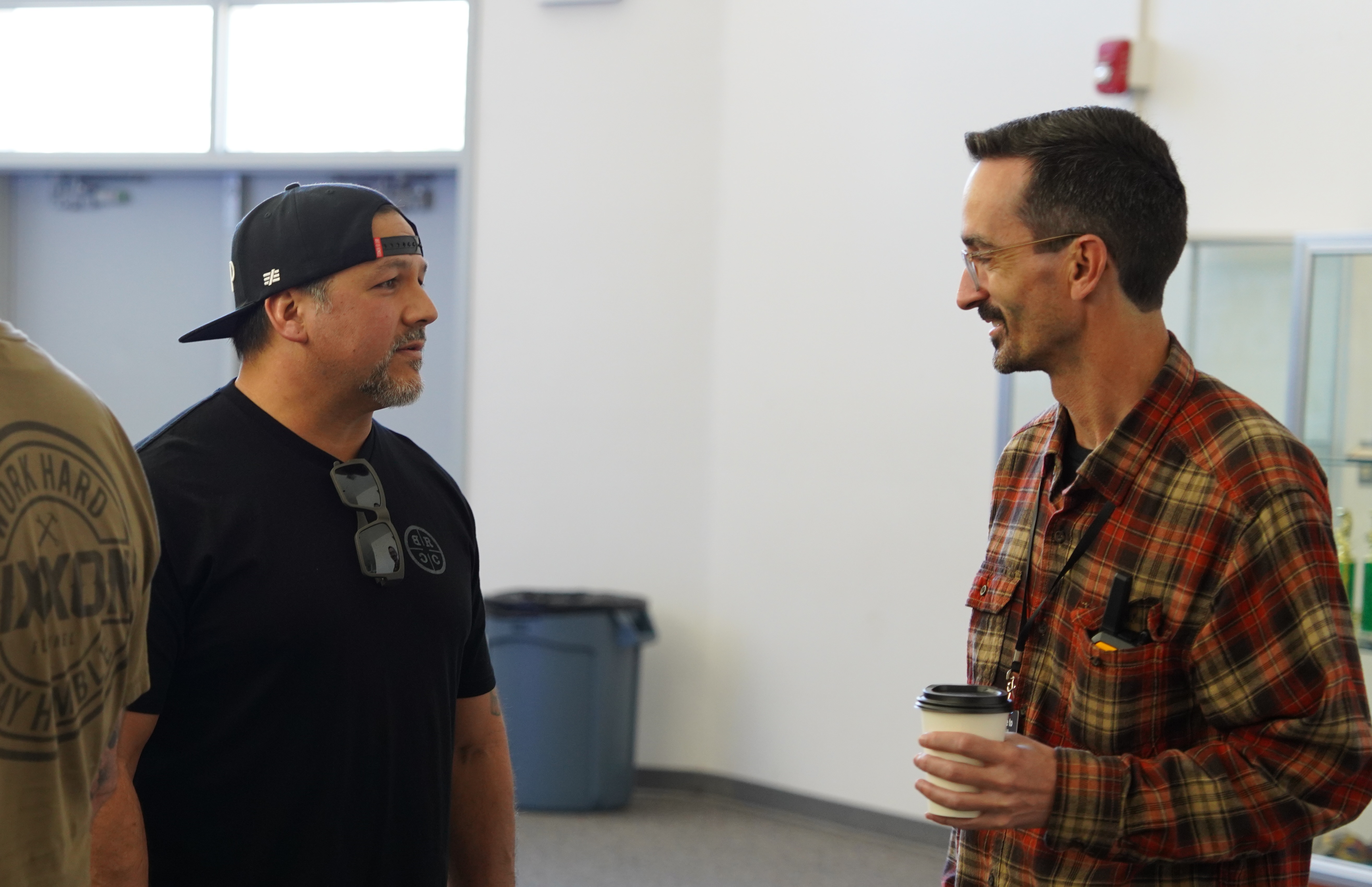 Two men talking indoors, one wearing a backward black cap and black t-shirt, the other in glasses and a red plaid shirt holding a coffee cup.