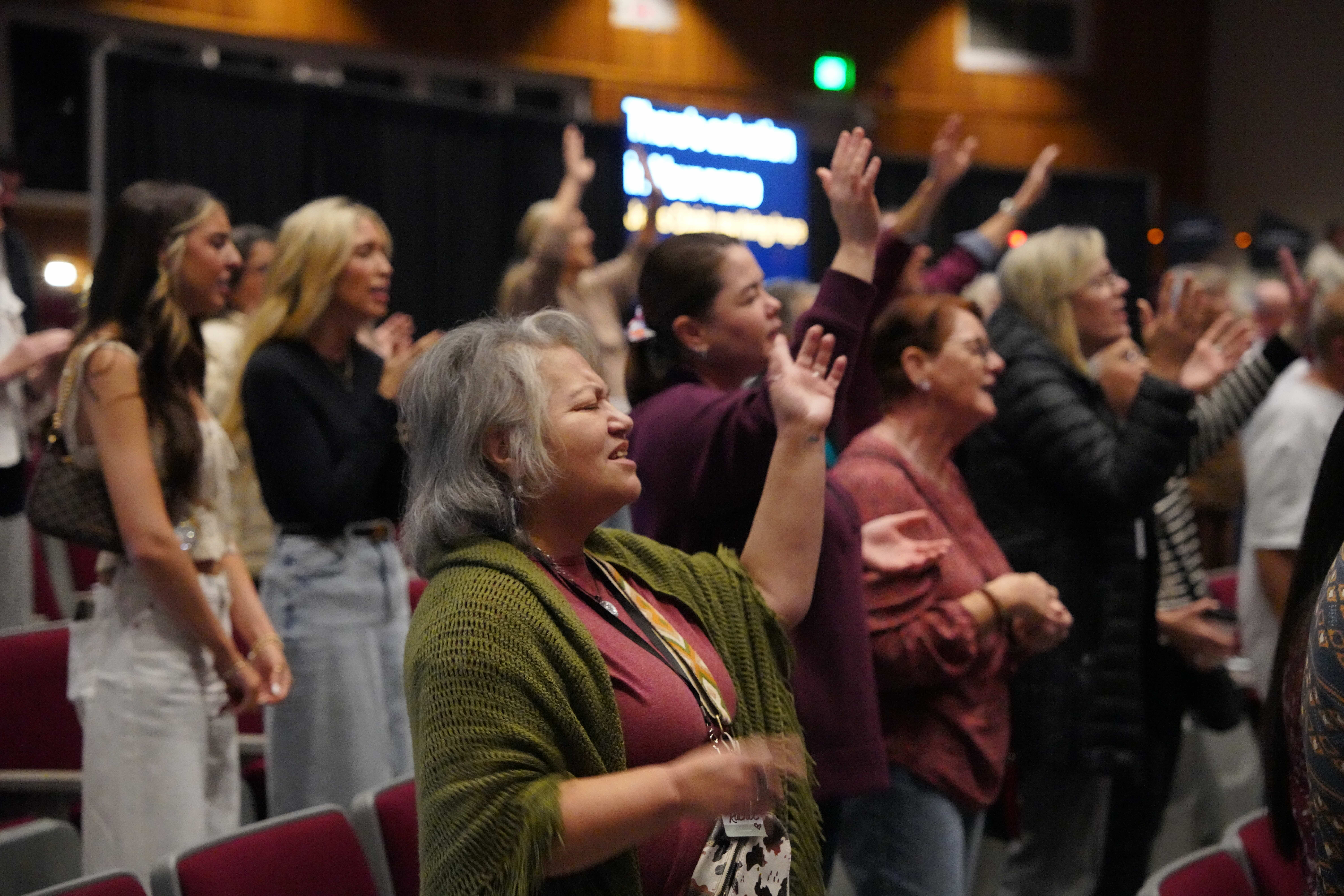 A diverse group of people with raised hands engaged in a worship service or concert in an indoor auditorium.