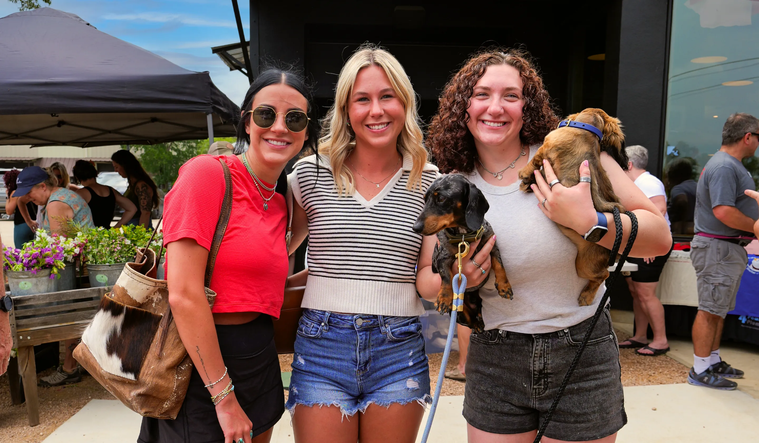 Three smiling women outdoors, one holding two small dogs, with market stalls and people behind them.