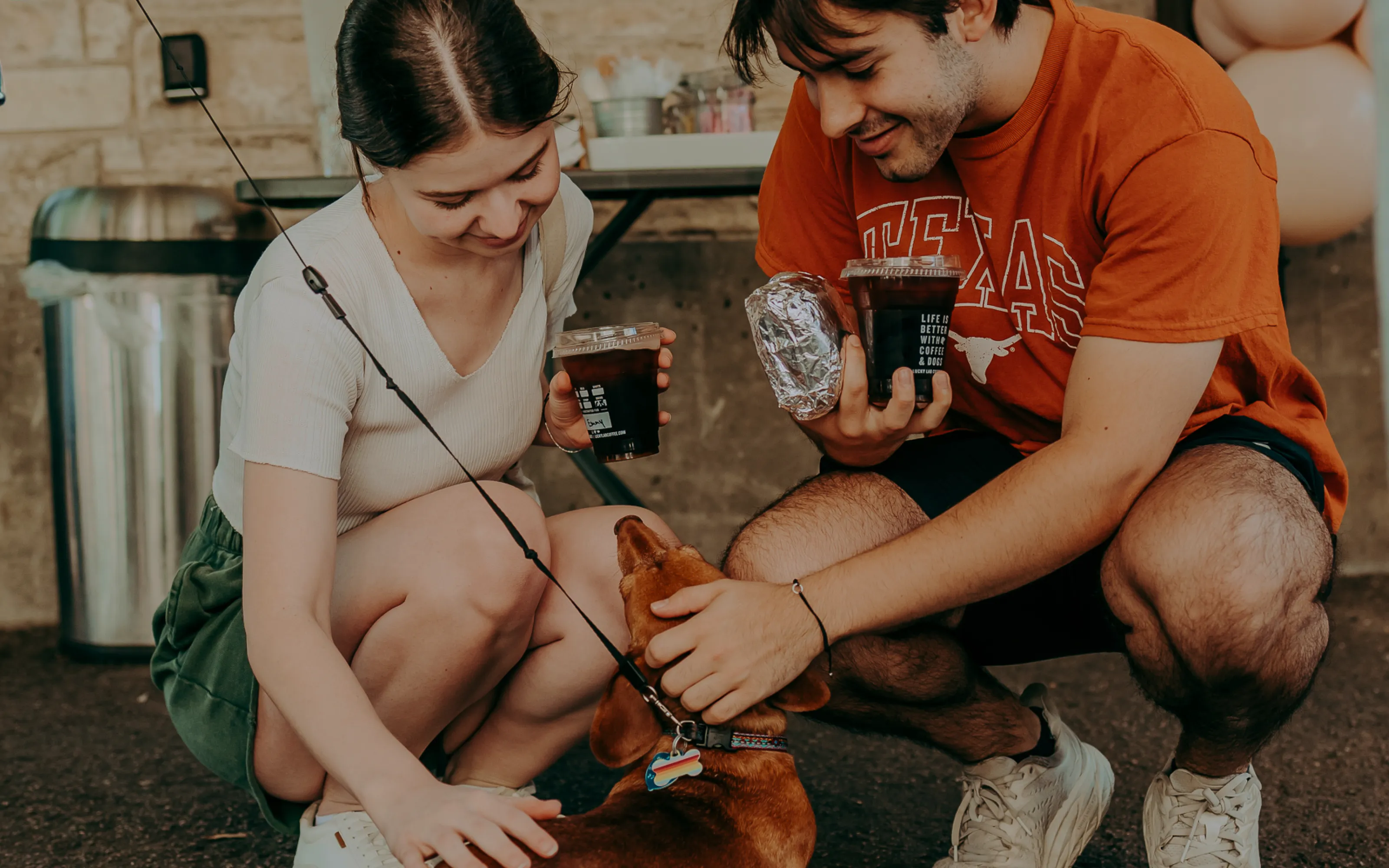 Young man and woman crouch to pet a brown dog while holding iced drinks and food wrapped in foil.