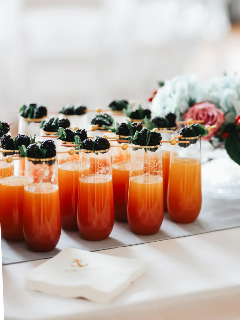 Rows of orange cocktails garnished with blackberries and mint leaves on wooden skewers at an event.