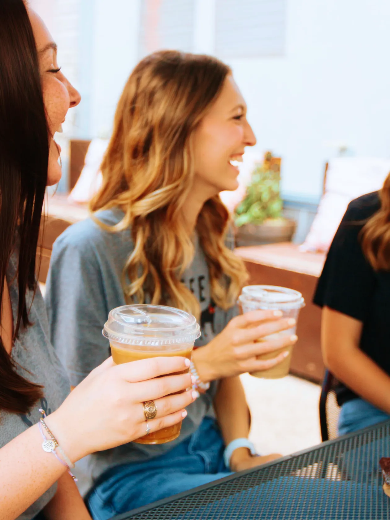 Two women smiling and holding plastic cups of iced coffee while sitting at a metal table outdoors.