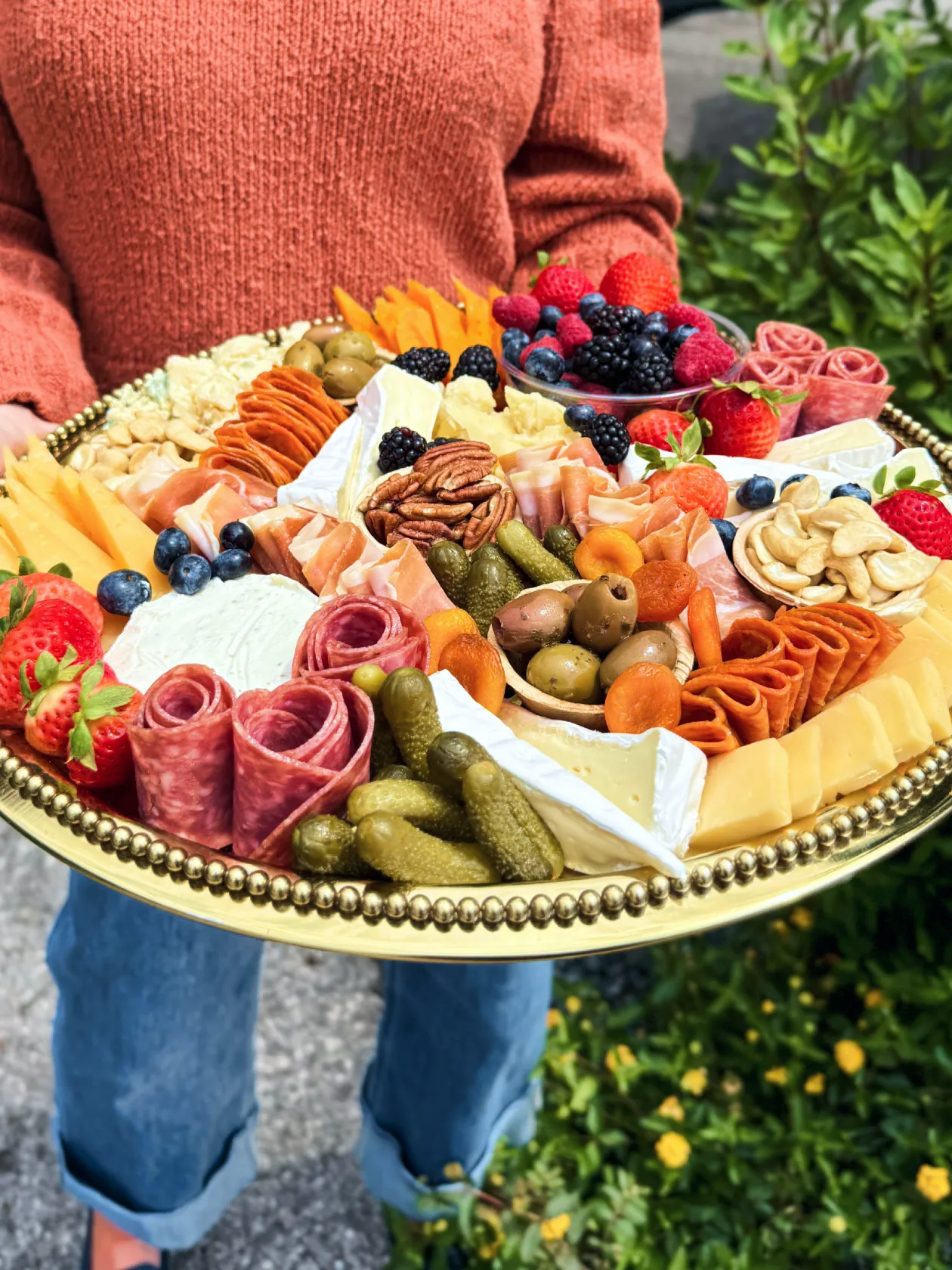 Person holding large platter with assorted cheeses, meats, fruits, nuts, olives, and pickles.