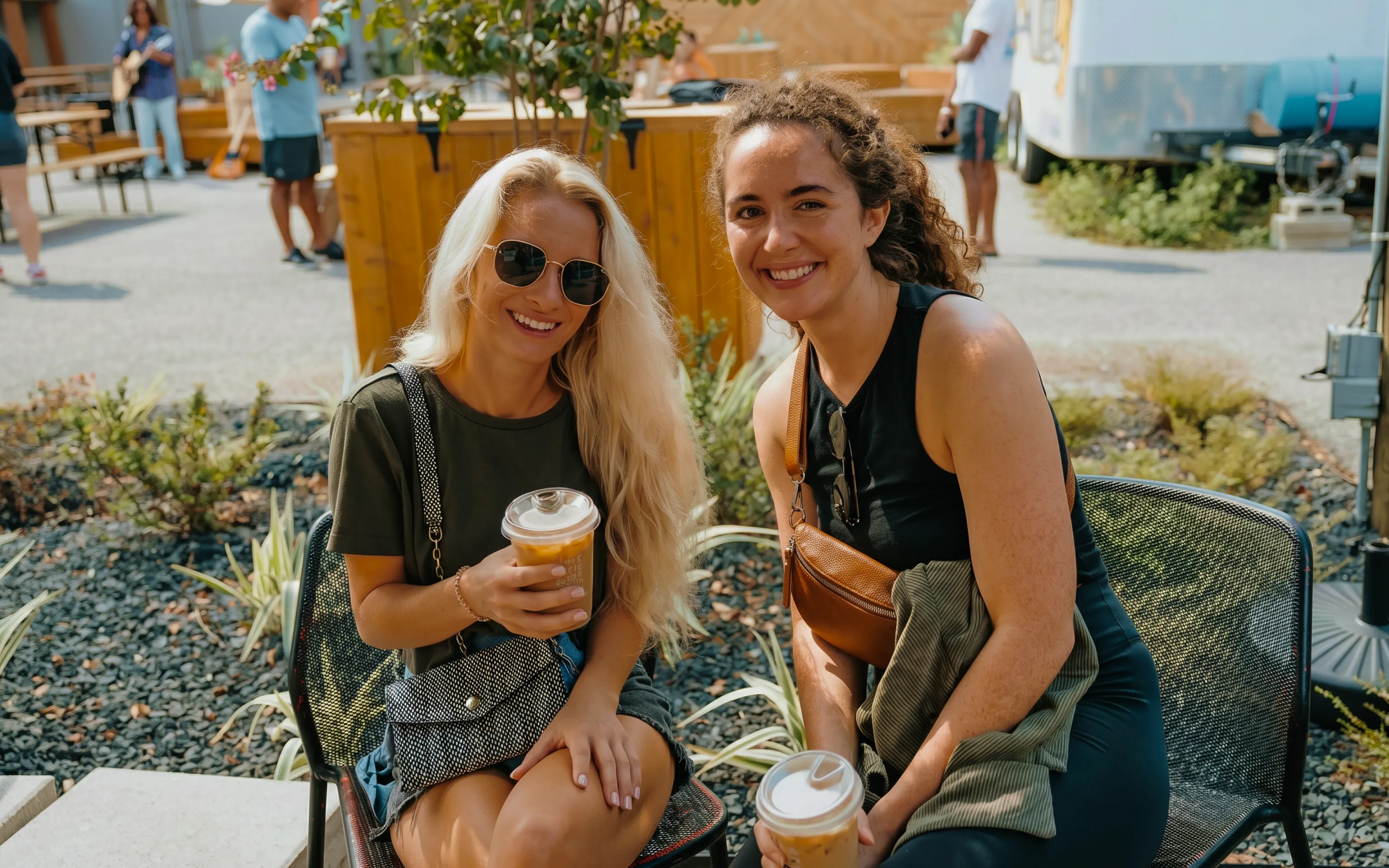 Two women sitting outside smiling, holding iced coffee drinks on a sunny day.