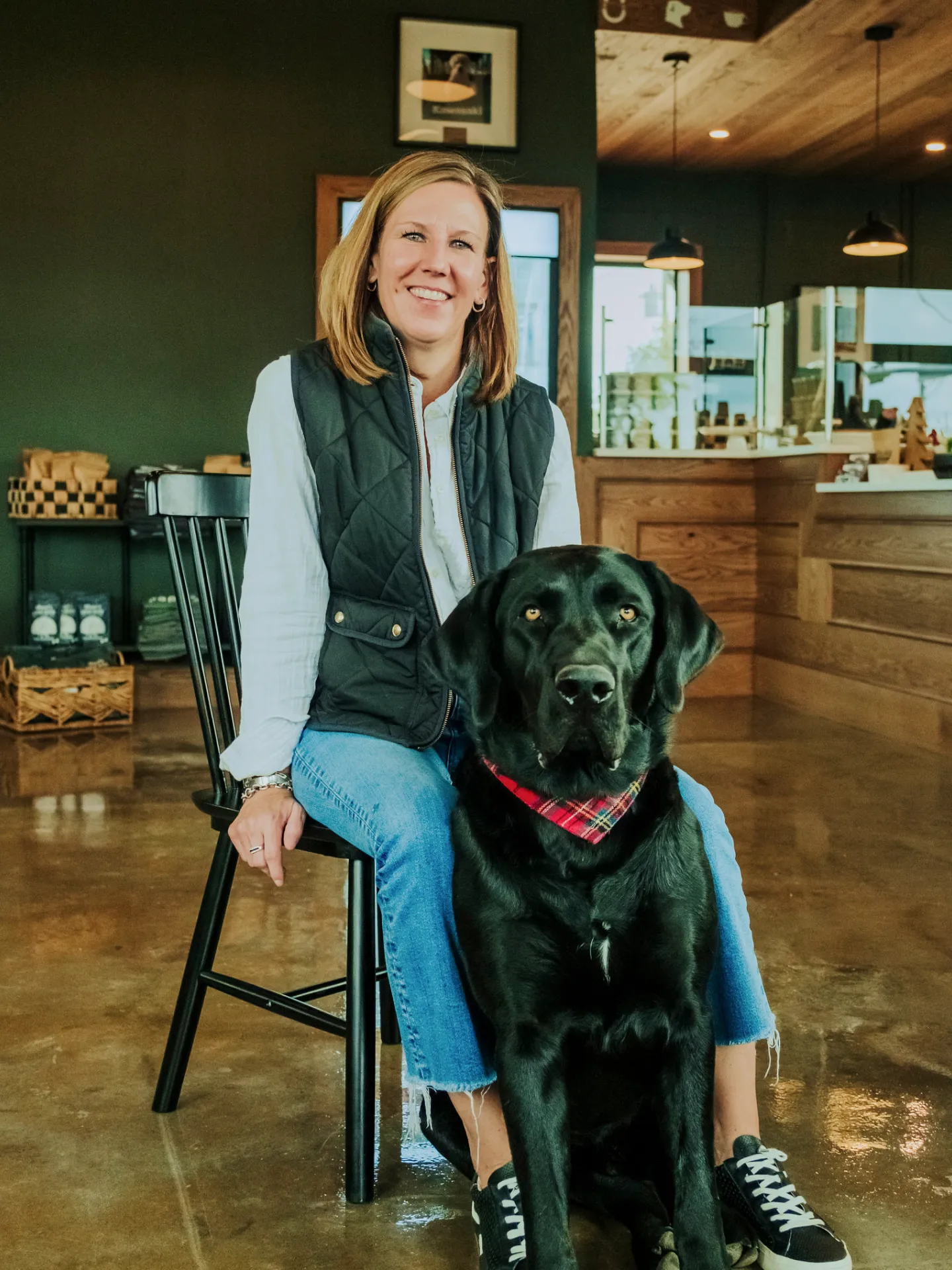 Smiling woman sitting on a chair indoors with a large black dog wearing a red plaid bandana.