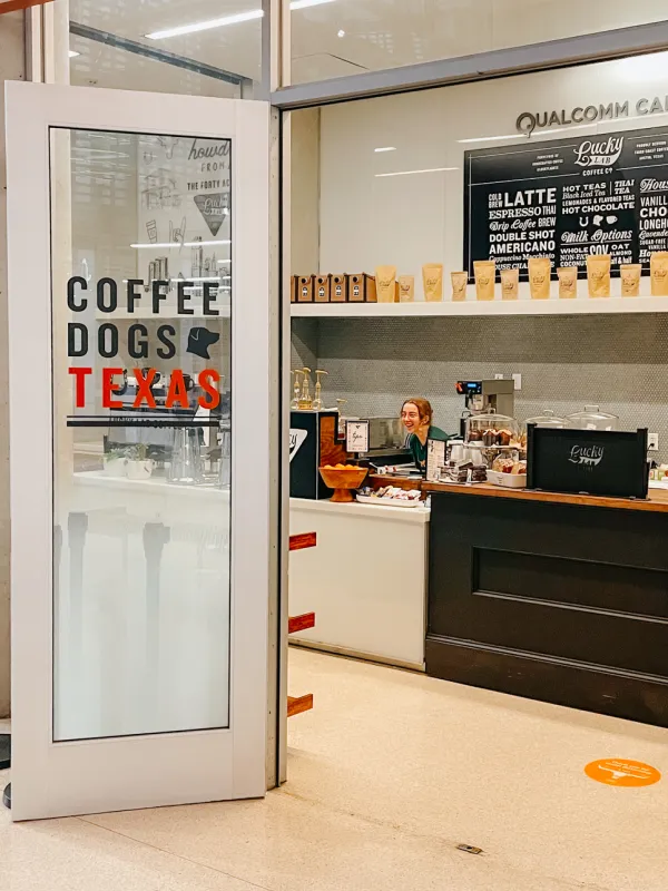 Open door of Coffee Dogs Texas café showing barista behind counter with coffee bags and menu board.