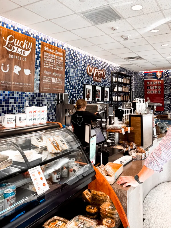 Barista prepares coffee behind a counter with pastries and blue tiled wall in Cappy's Place cafe.