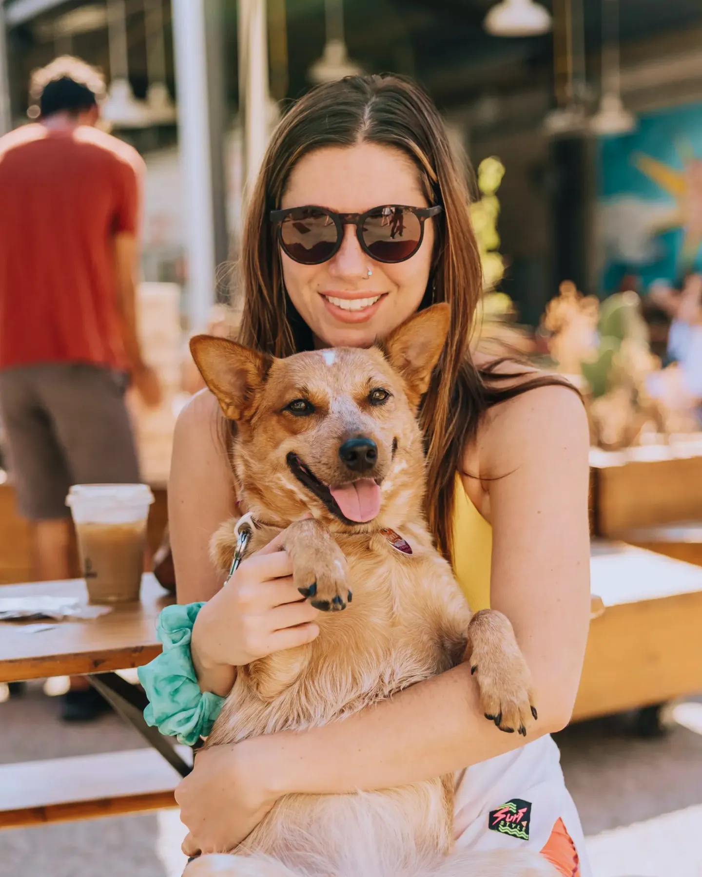 Black Labrador retriever sitting outdoors with tongue out, leash held by a seated woman in background.