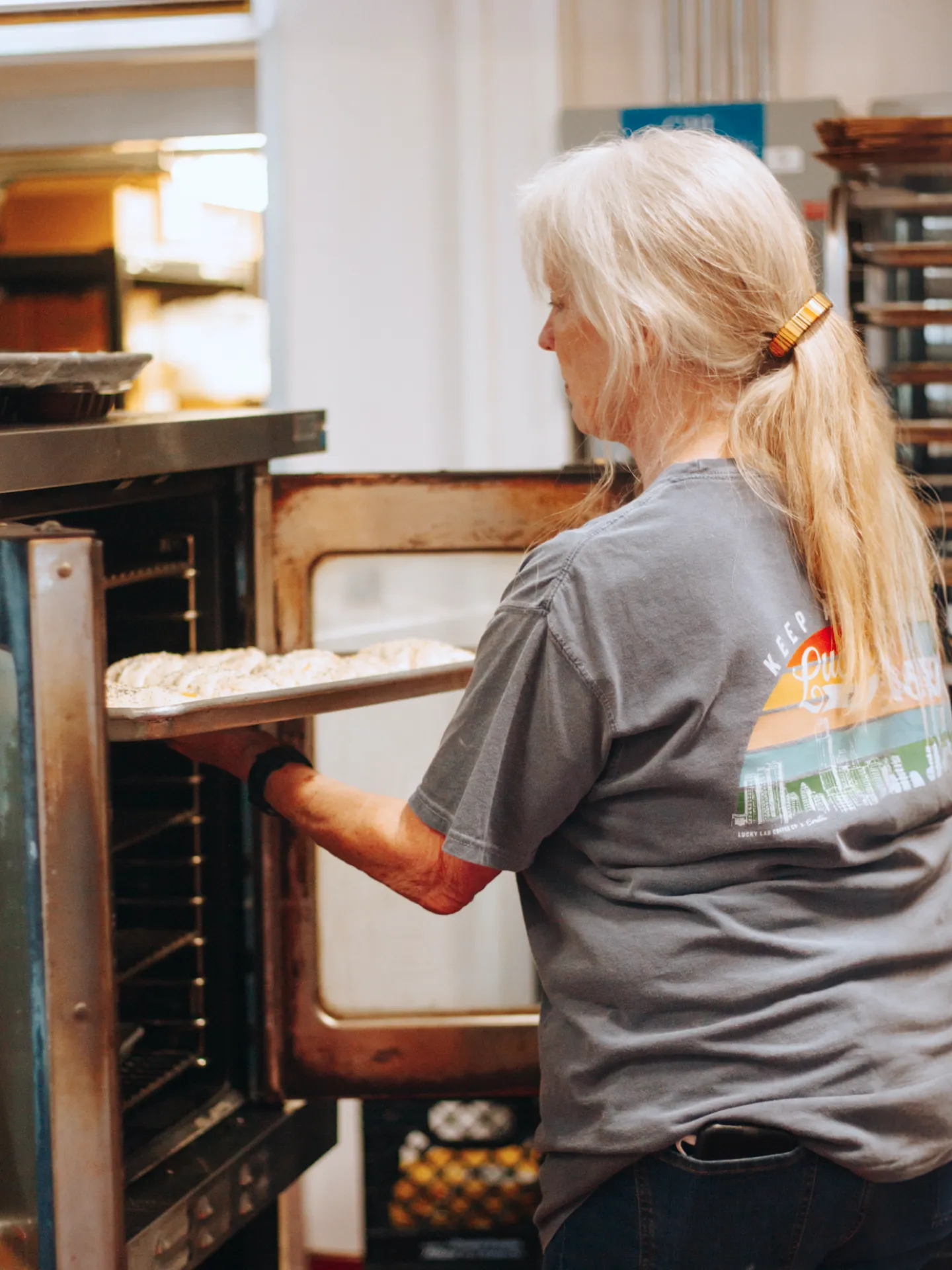 Woman placing a tray of dough or pastries into an open oven in a bakery kitchen.
