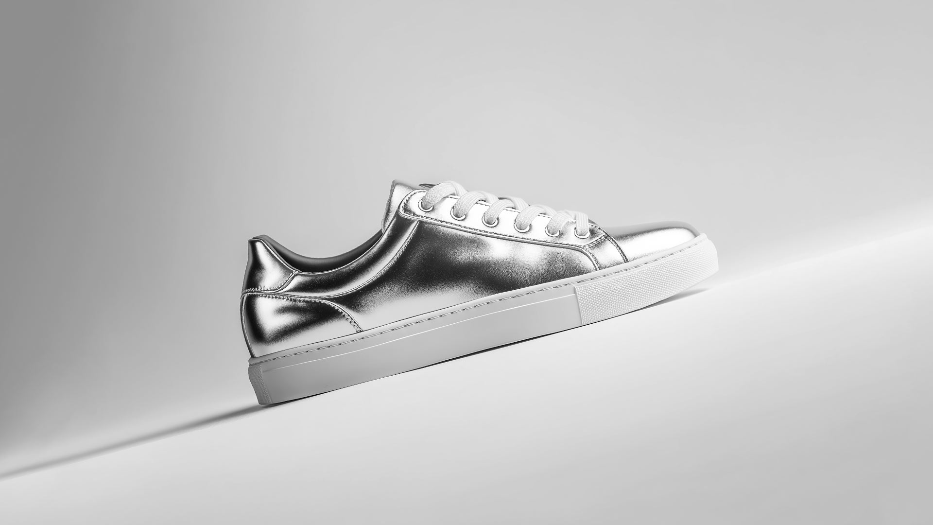 Side view (rotated) of a minimalist metallic silver low-top sneaker with white laces and a white rubber sole, floating against a light gray background.