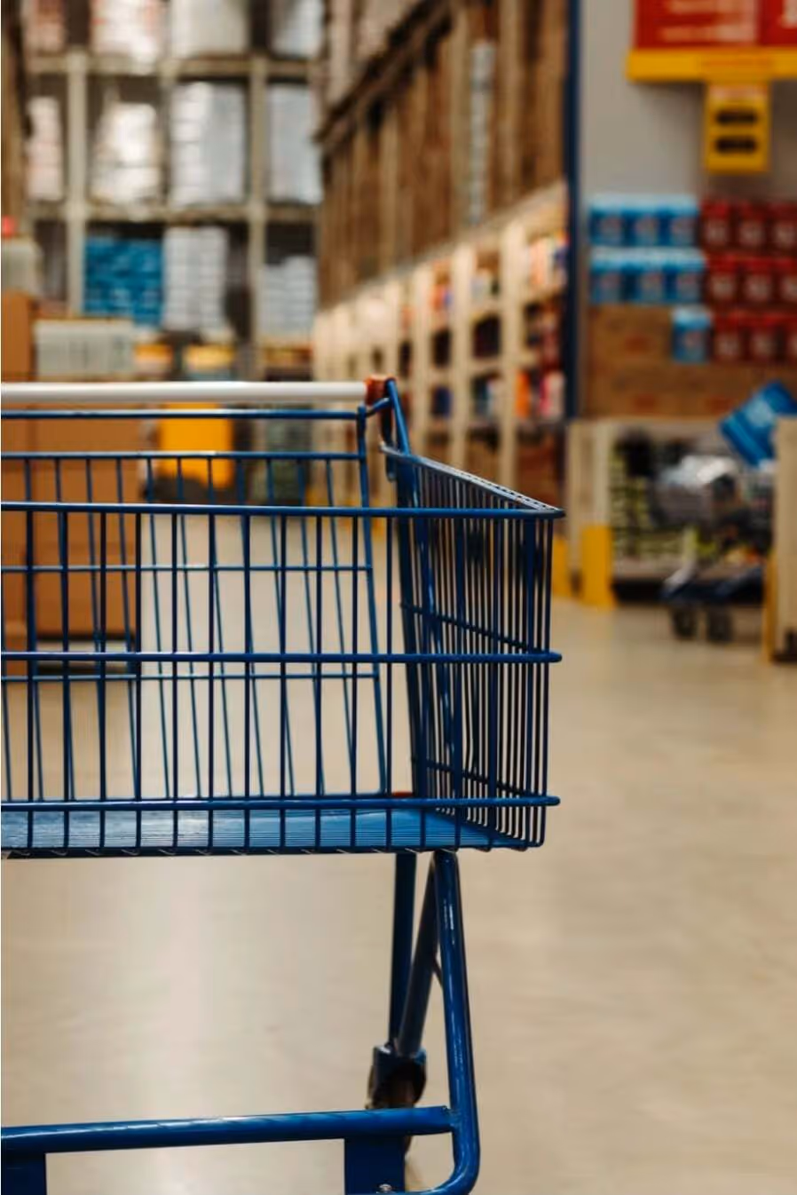 Empty blue shopping cart in the aisle of a warehouse-style store with shelves stocked in the background.