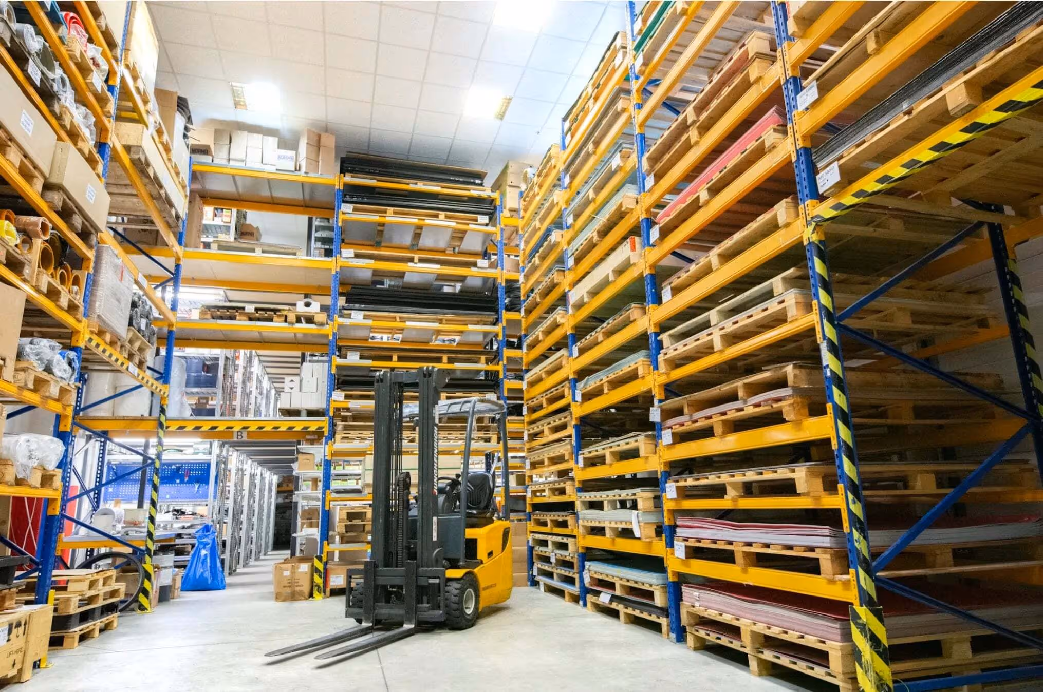 Warehouse aisle with tall industrial shelving filled with pallets and boxes, and a yellow forklift parked in the center.