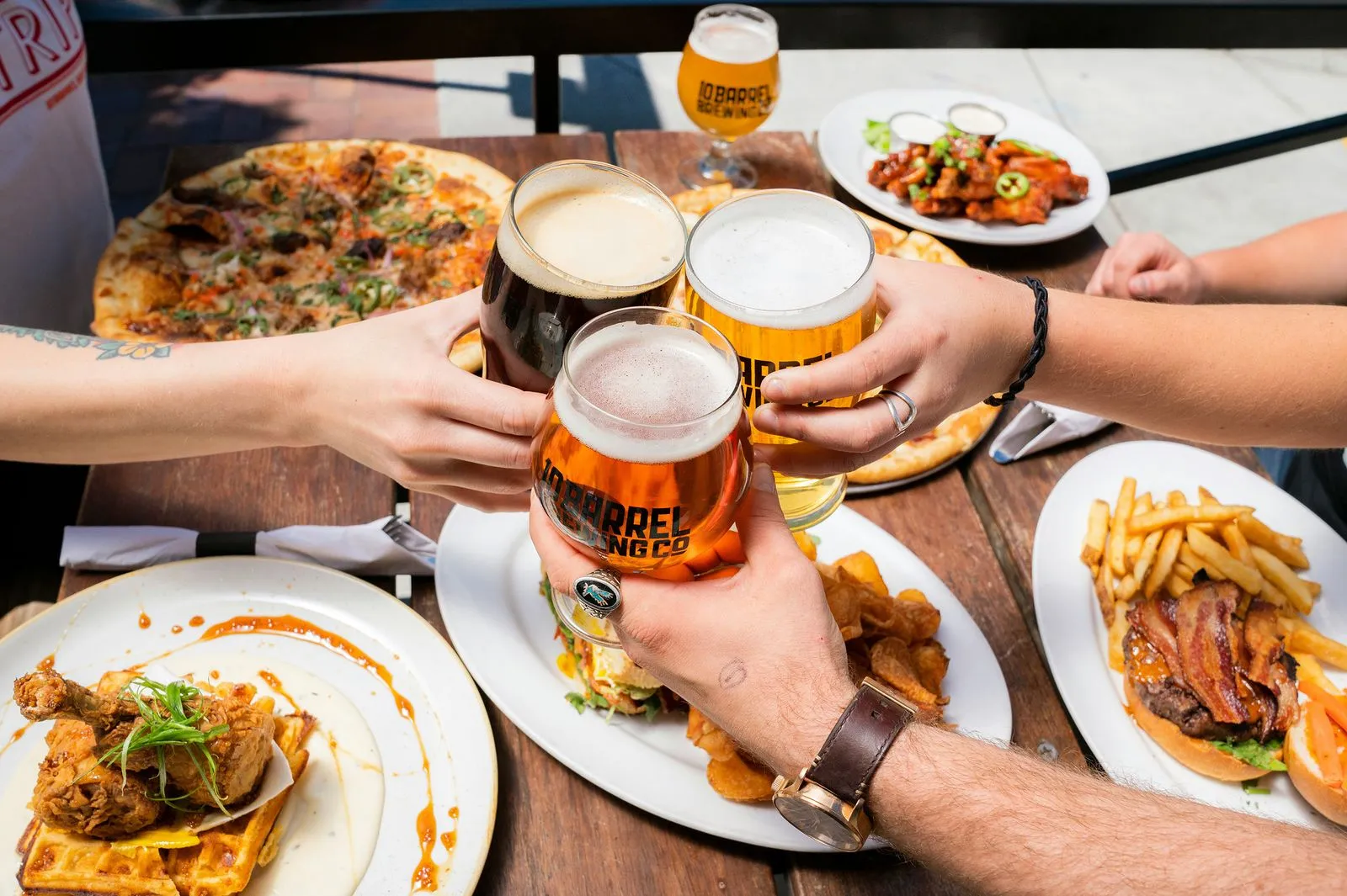 Four hands clinking beer glasses over table of food