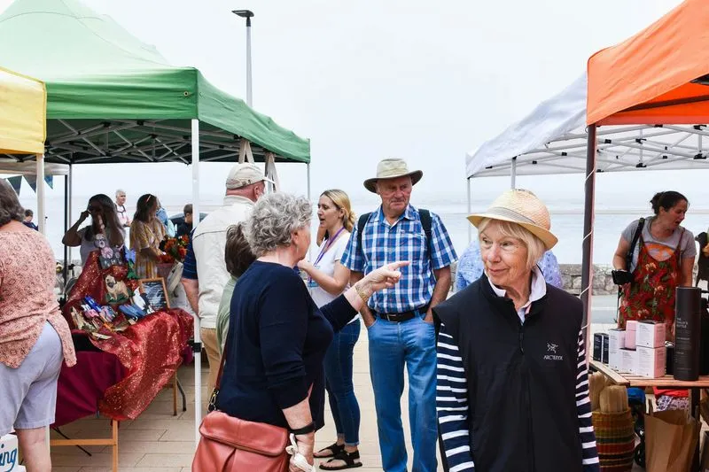 People enjoying a lively outdoor market by the sea, chatting and browsing stalls under colorful tents on a cloudy day, creating a vibrant atmosphere.
