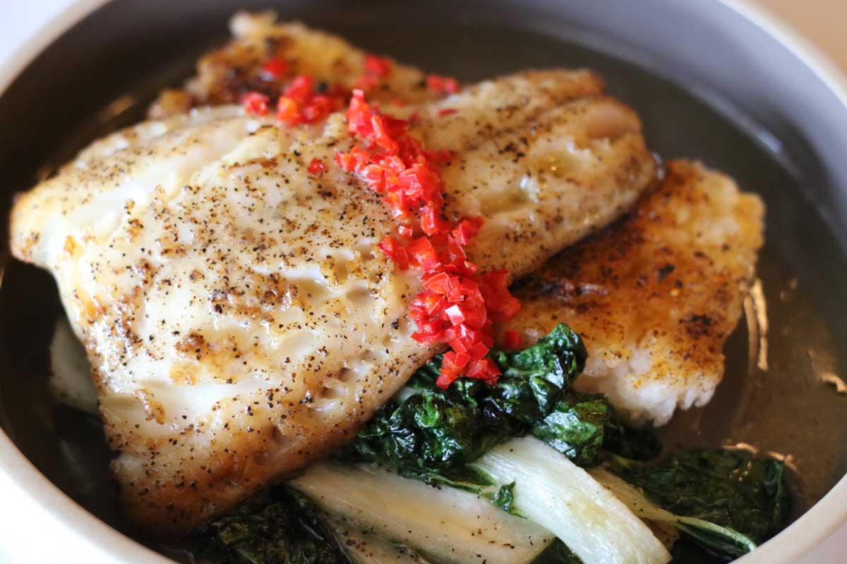 Pan-seared white fish fillets seasoned with black pepper and topped with chopped red peppers served over sautéed greens in a round bowl.