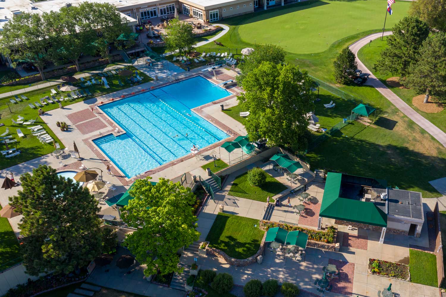 Aerial view of an outdoor swimming pool with adjacent patio areas, lounge chairs, shaded seating, and a surrounding golf course green.