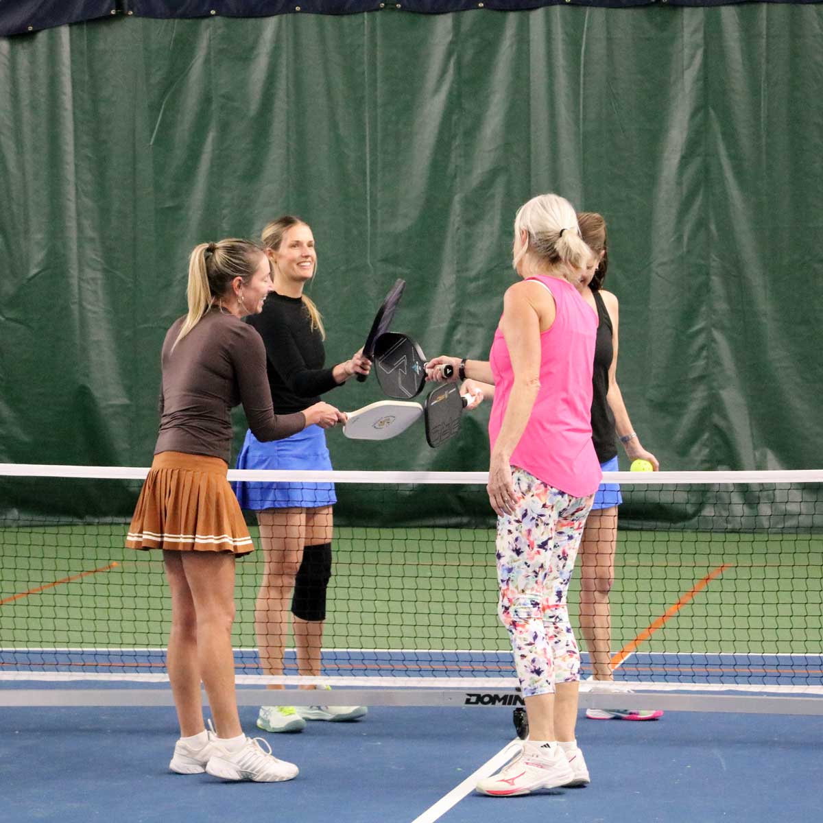 Four women standing on a pickleball court with paddles, smiling and interacting near the net.