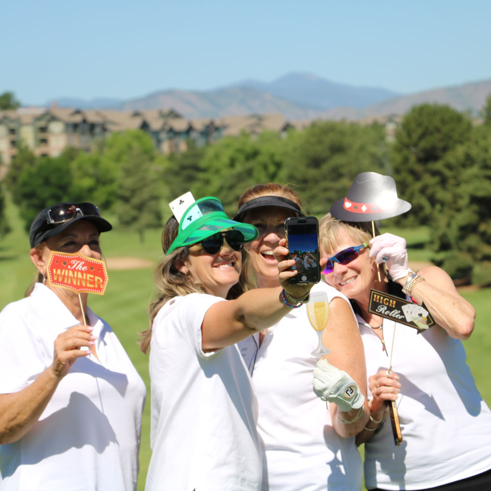 Four women on a golf course smiling and holding playful signs and props, taking a selfie together.