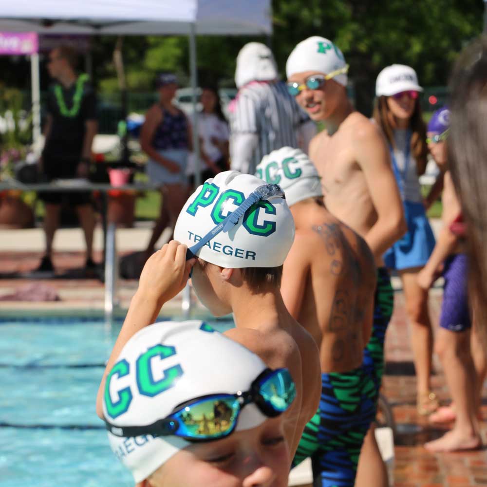 Swimmers wearing white PCC swim caps and green-and-black swim jammers preparing by the poolside on a sunny day.
