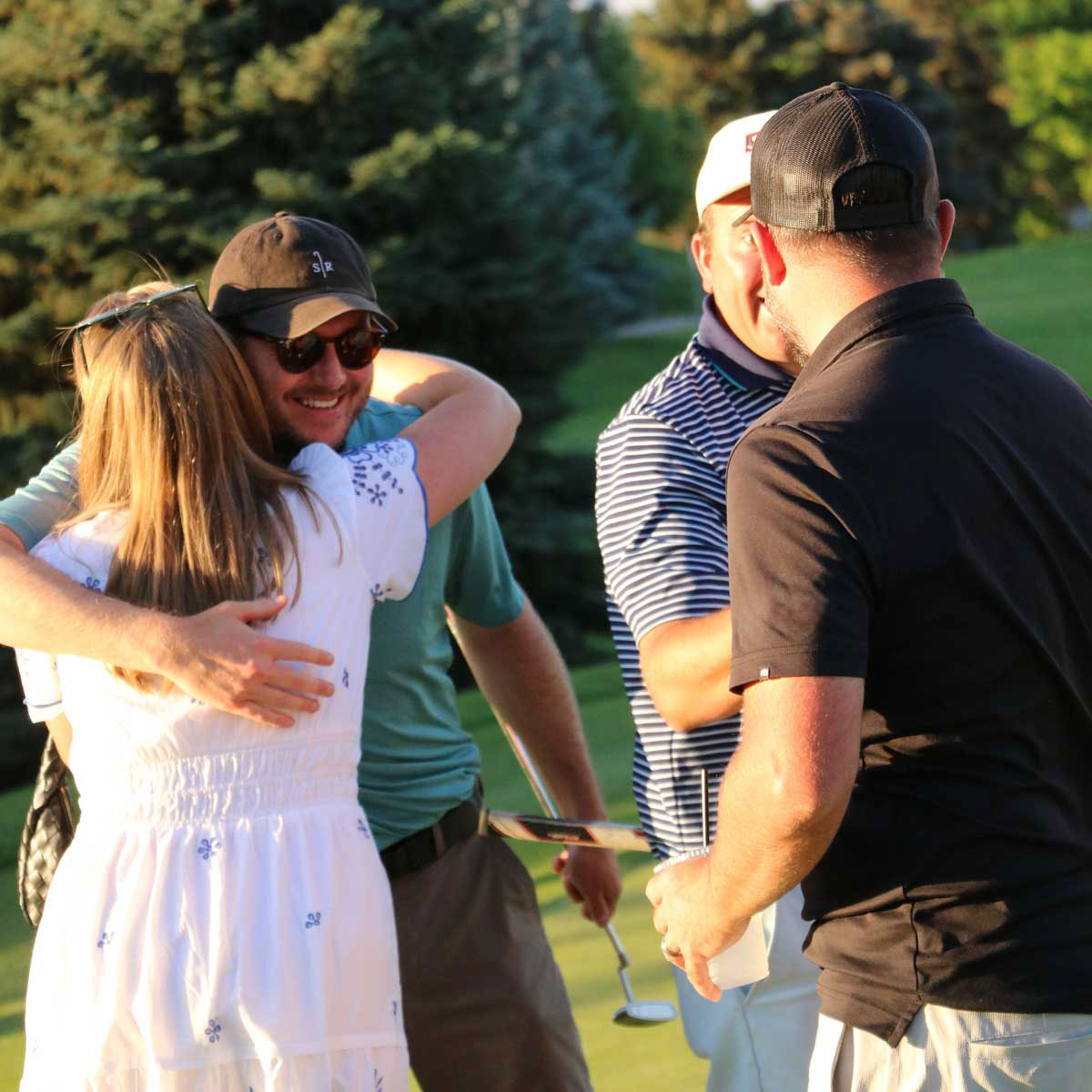 Two men and a woman outdoors on a golf course, with one man wearing sunglasses and a cap hugging the woman, and two other men smiling and talking nearby.
