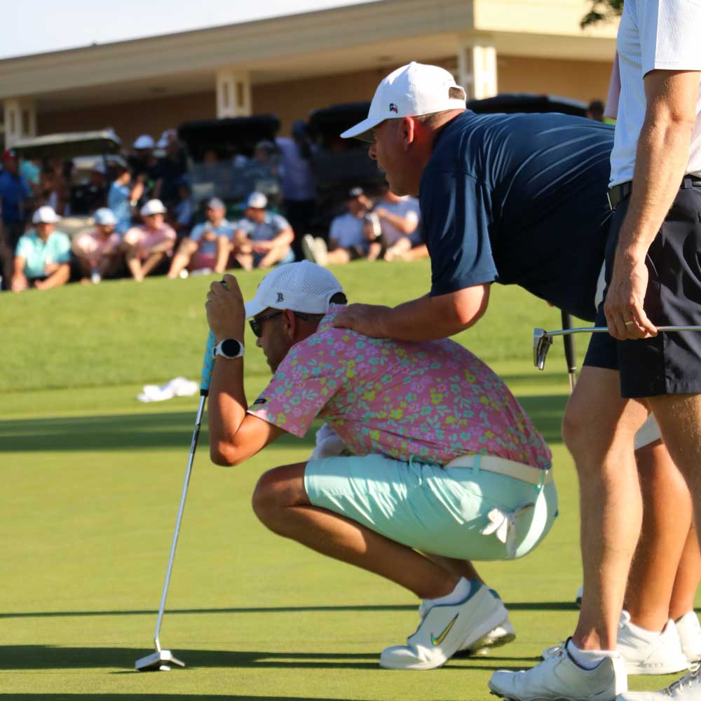 Two male golfers on green, one squatting in a pink floral shirt and mint shorts lining up a putt, the other in navy shirt with hand on squatter's shoulder.