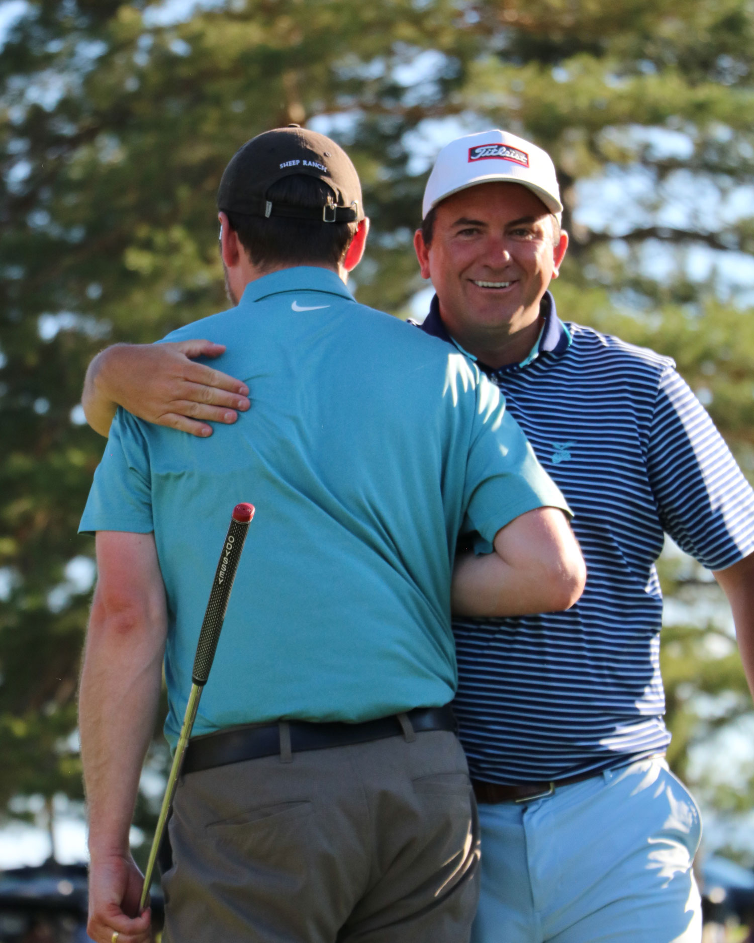 Two men in golf attire embracing outdoors, one holding a golf club and smiling at the camera.