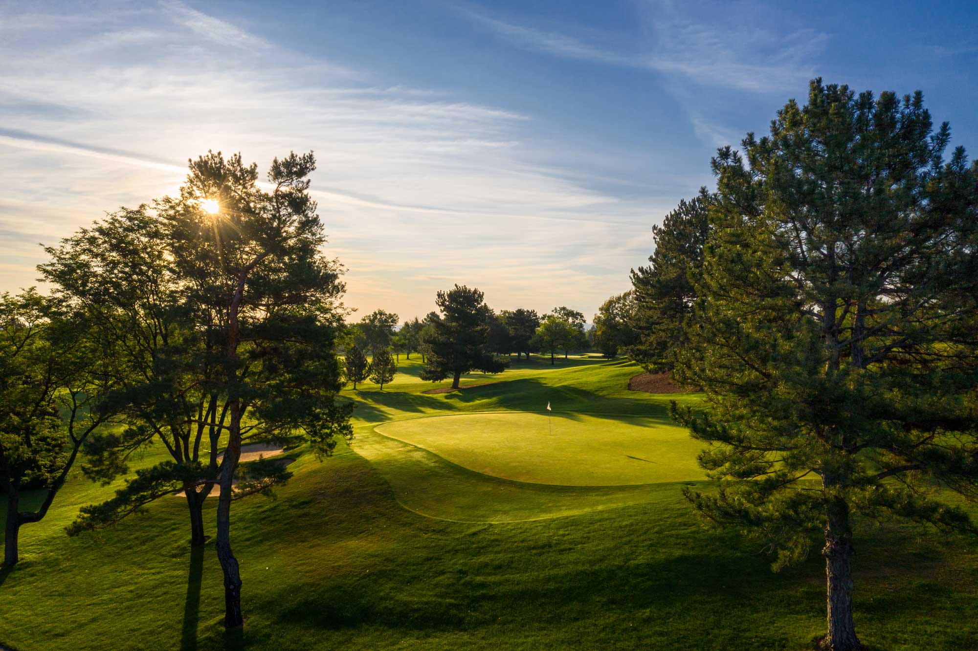 Sunlight shining through trees over a golf green with a flagstick on a clear day.