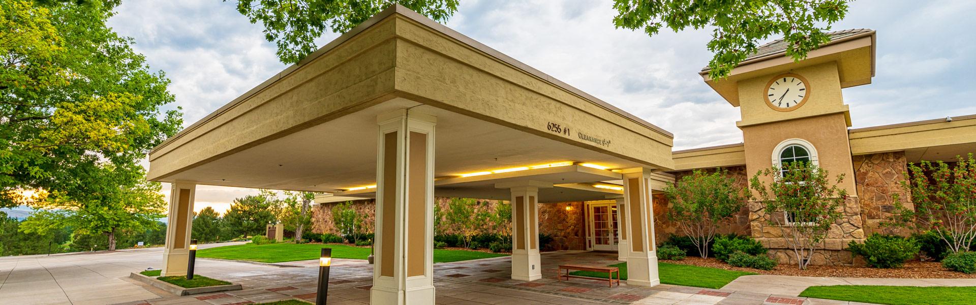 Building entrance with covered driveway supported by columns, clock tower, and surrounding greenery under a cloudy sky.
