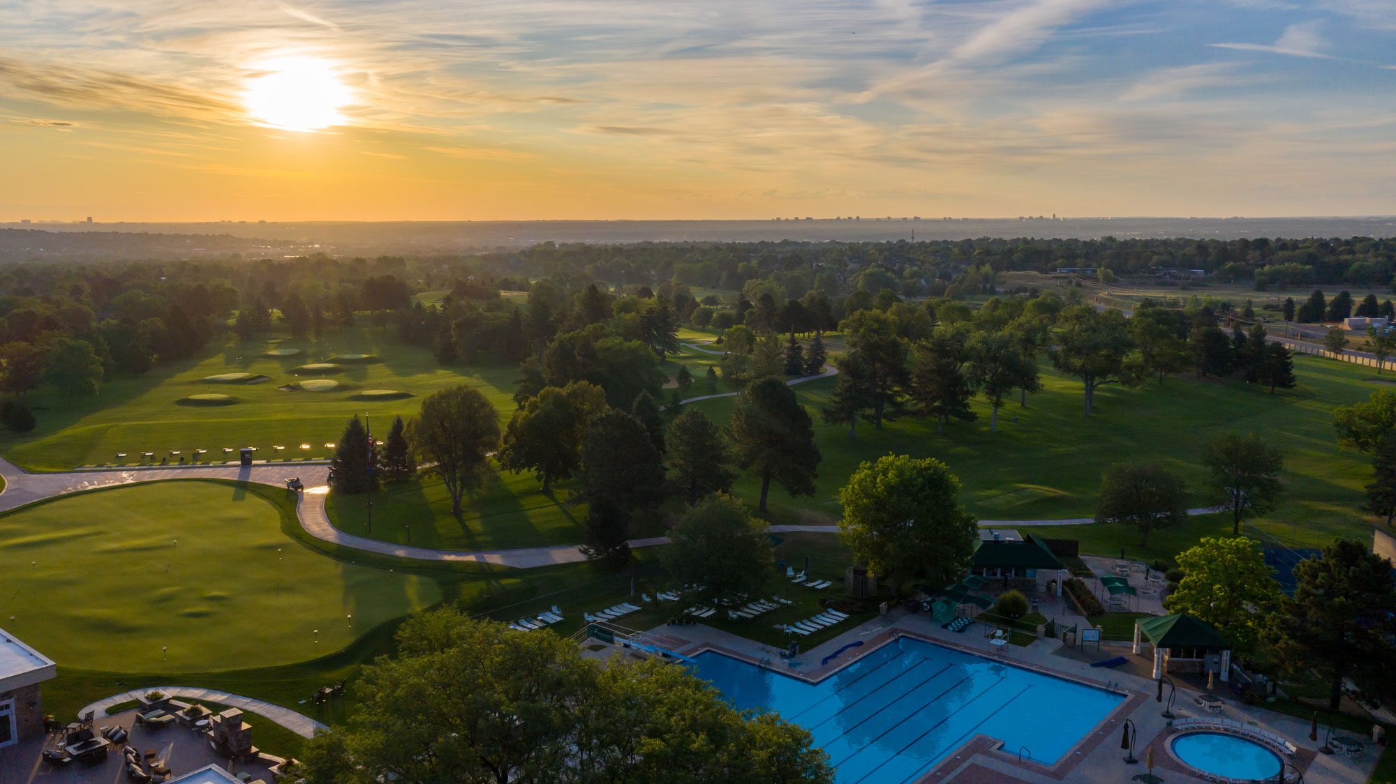 Aerial view of a golf course and outdoor swimming pool at sunset with trees and a distant city skyline.