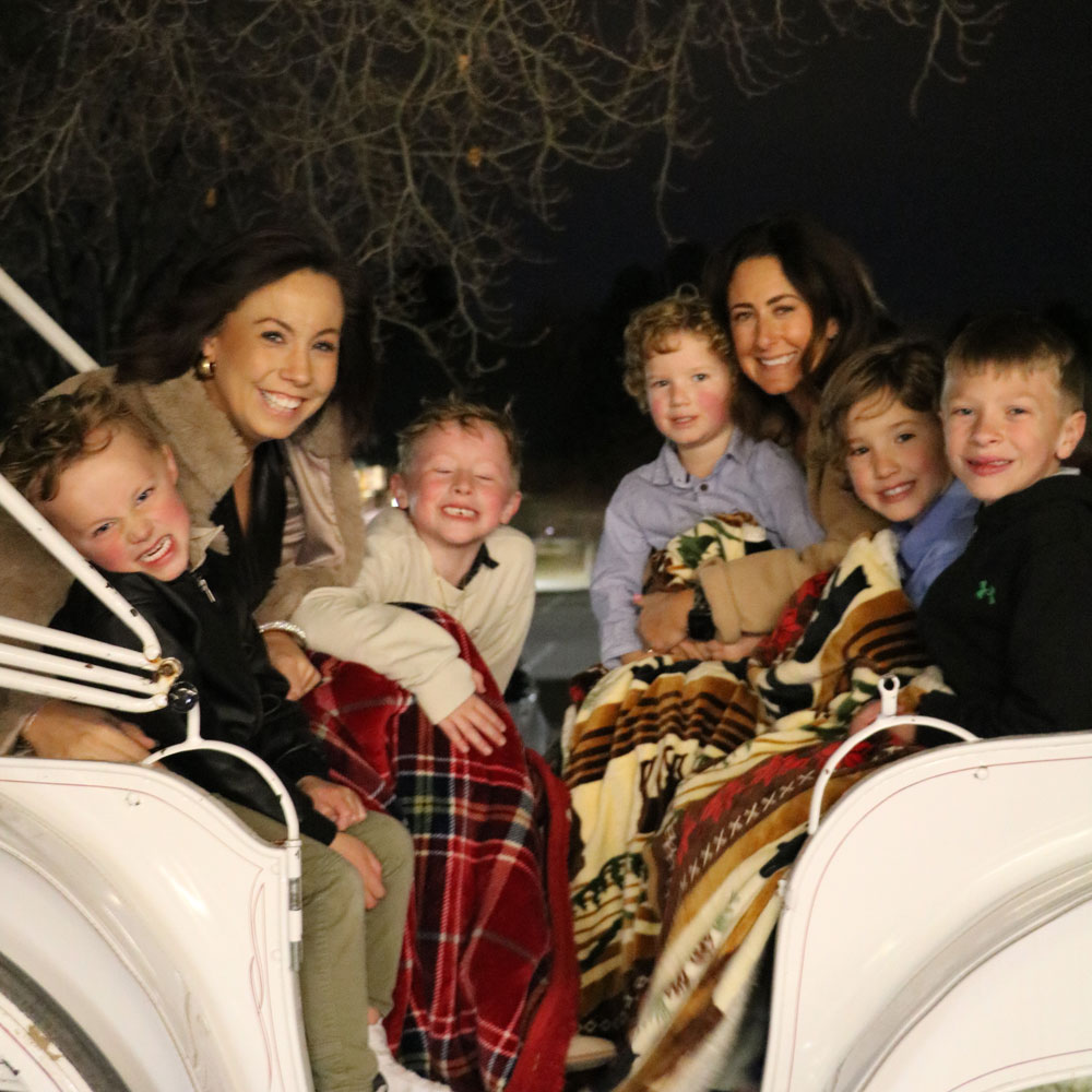 Two women and five children sitting closely together on a white carriage at night, wrapped in colorful blankets and smiling.