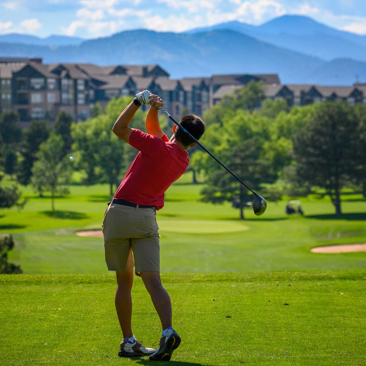 Golfer in a red shirt and beige shorts finishing a swing on a green golf course with trees and buildings in the background.