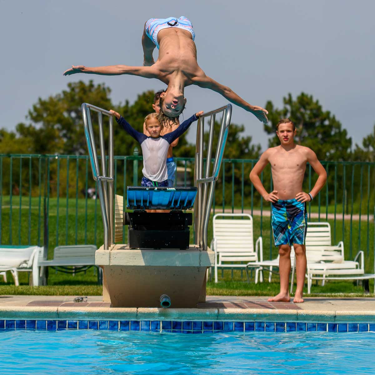 Boy in blue striped swim trunks executing a backflip off the diving board while other children wait nearby at a pool.