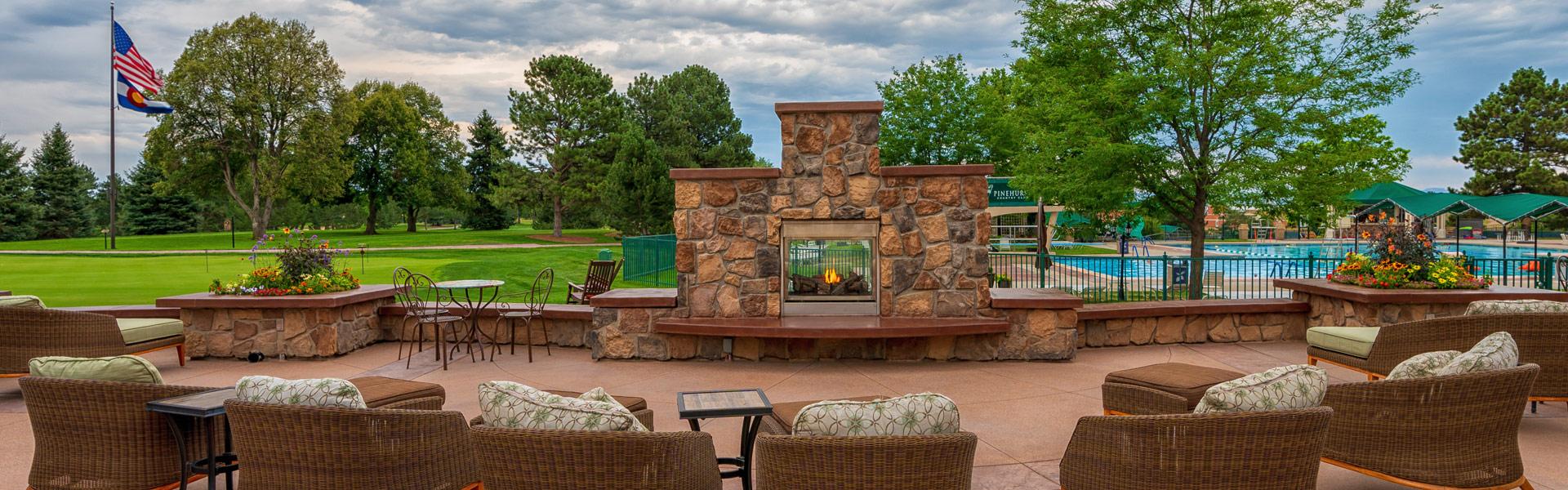 Outdoor patio with wicker seating and cushions facing a stone fireplace, with a golf course and swimming pool in the background.