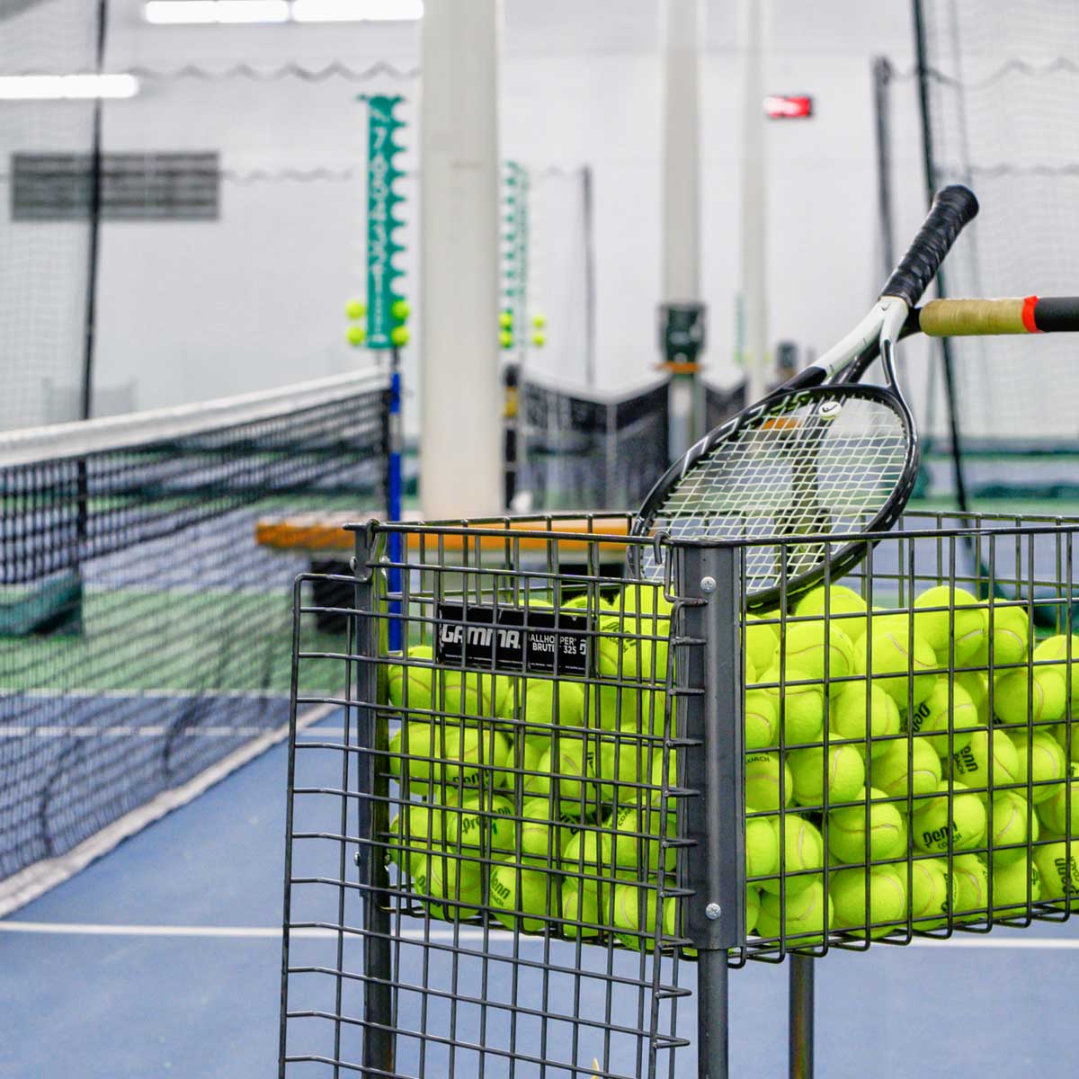Wire basket filled with yellow tennis balls and a tennis racket on an indoor tennis court.