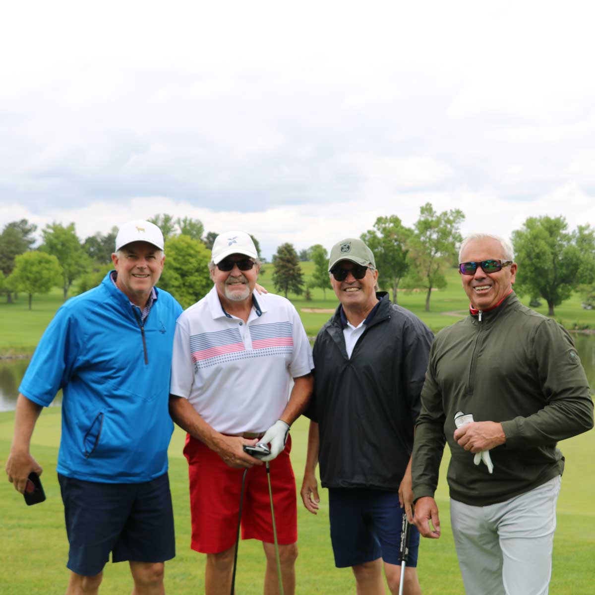 Four men smiling on a golf course, dressed in casual golf attire and holding golf clubs.