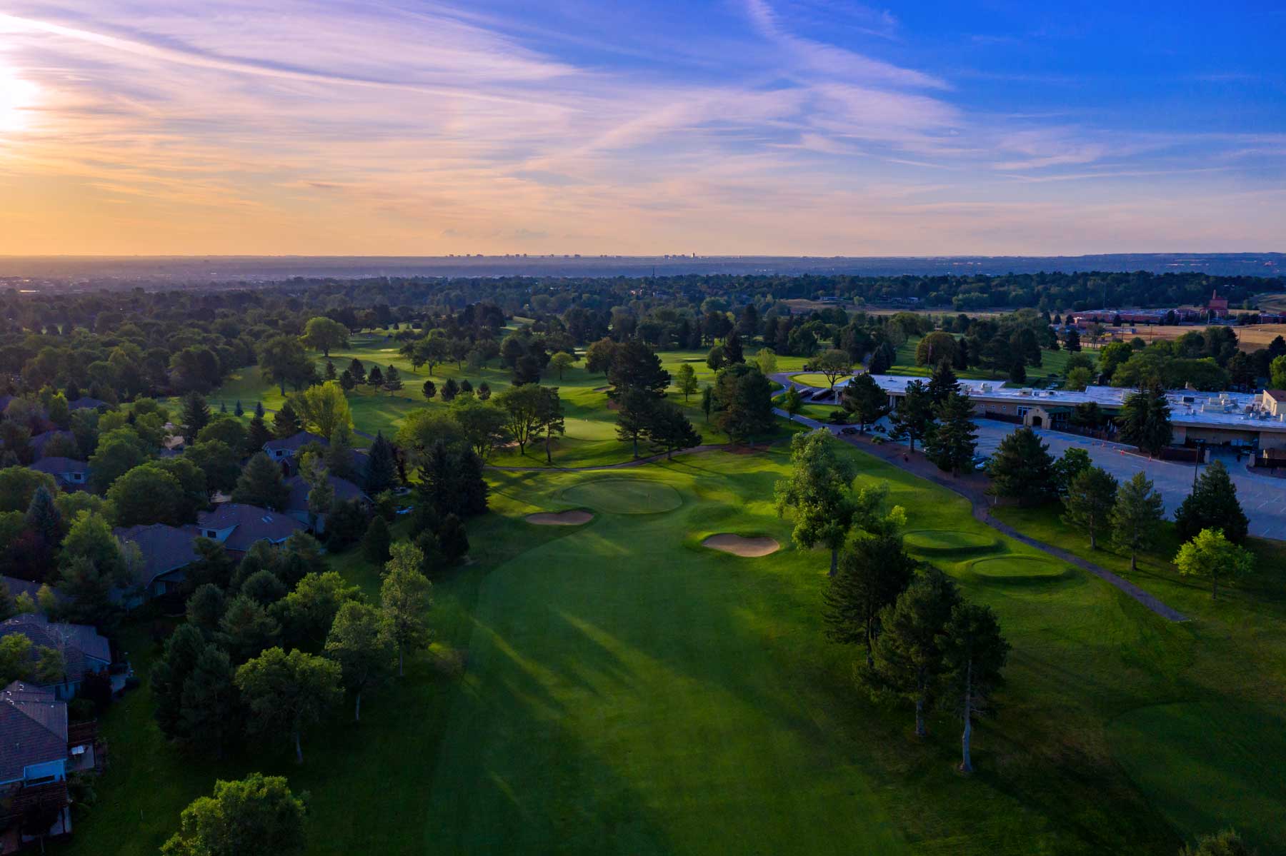 Aerial view of a green golf course with sand bunkers, surrounded by trees and residential houses under a colorful sunset sky.