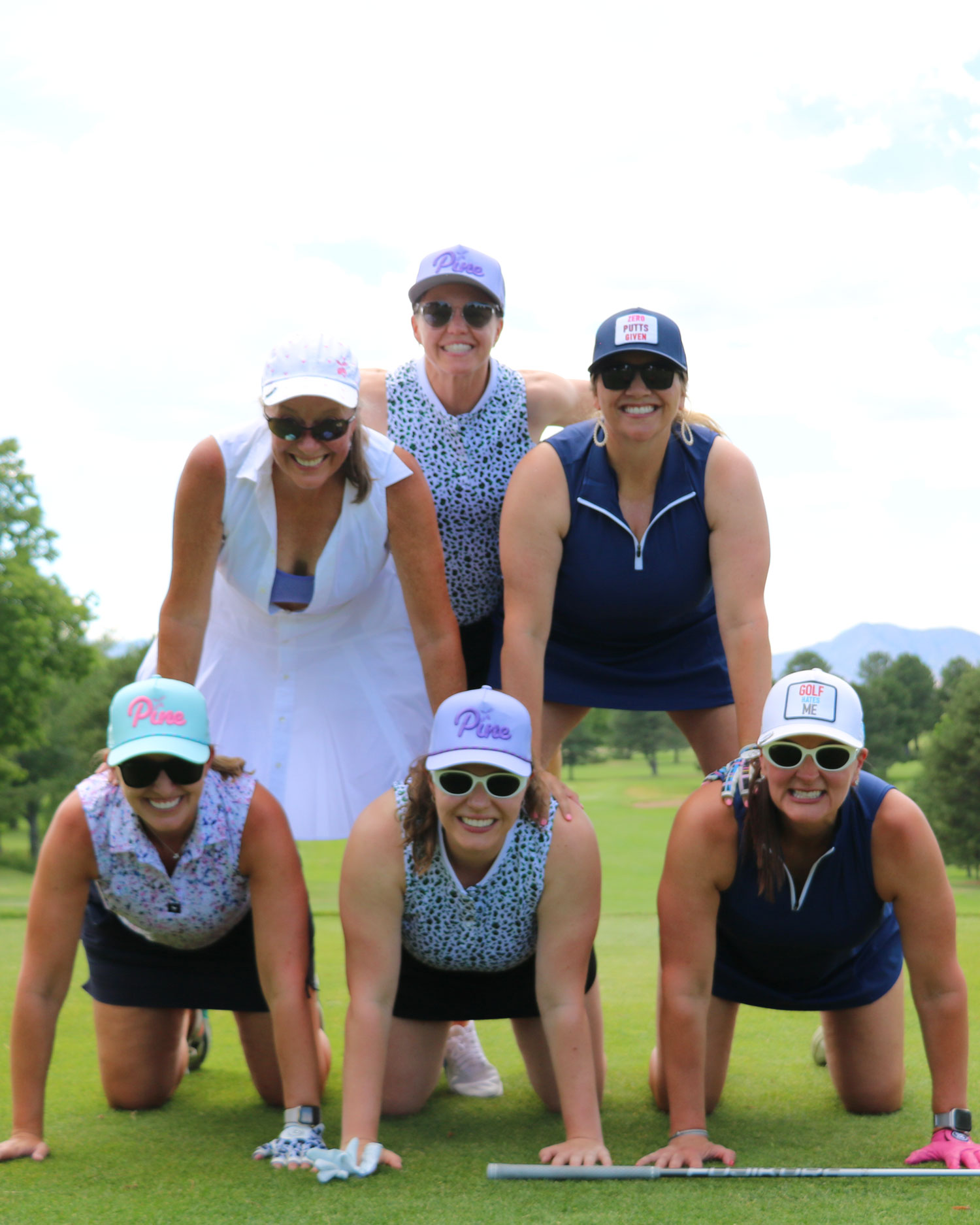 Six women in colorful golf attire forming a human pyramid on a golf course, smiling at the camera.
