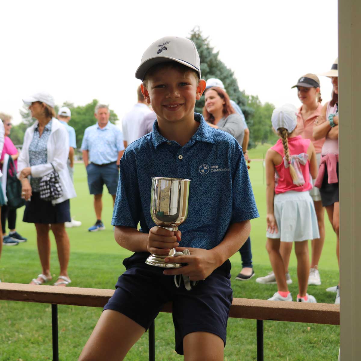 Young boy in a blue BMW Championship shirt and gray cap holding a silver trophy cup while sitting on a wooden railing with people and a golf course in the background.