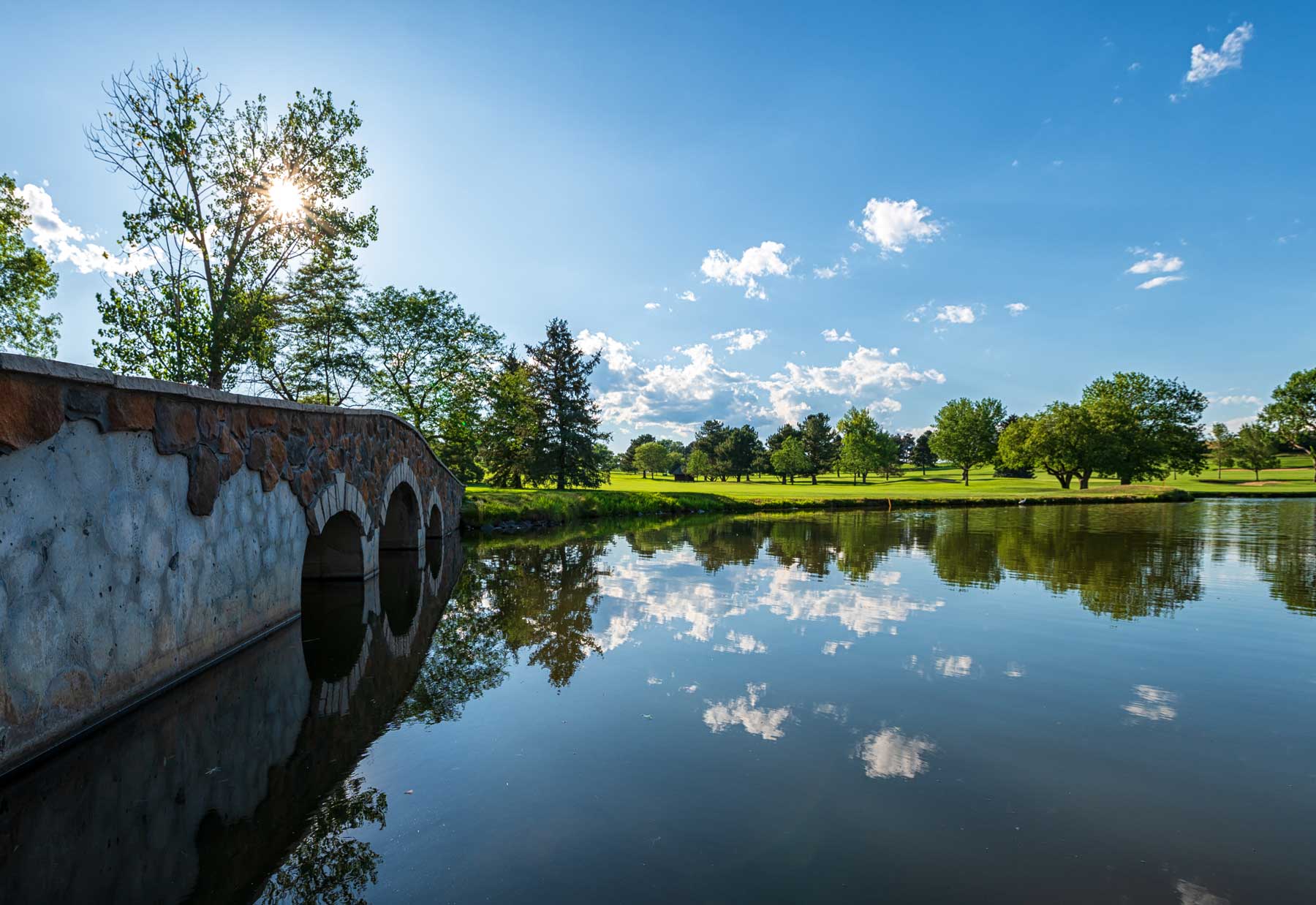 Stone bridge over a calm pond reflecting blue sky, clouds, and surrounding trees on a sunny day.