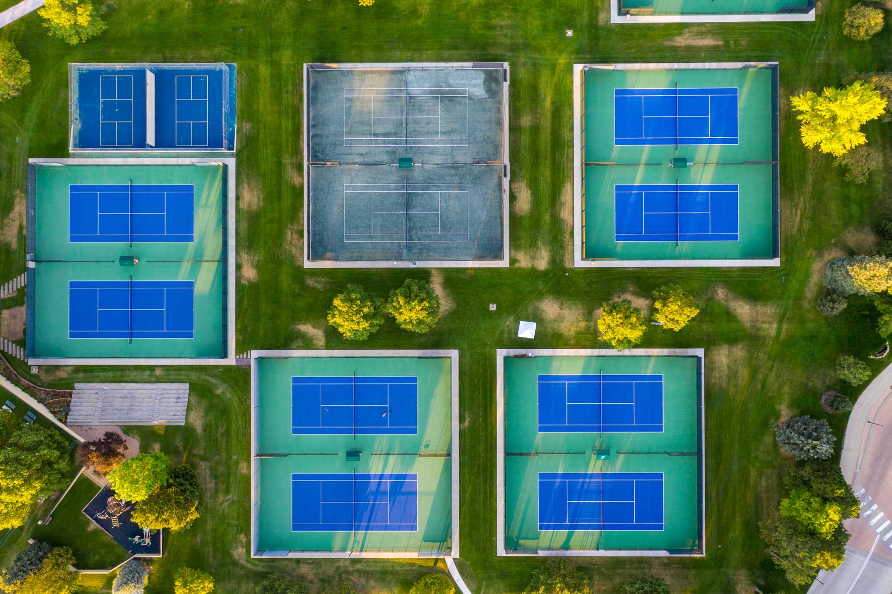 Aerial view of multiple outdoor tennis courts with blue playing surfaces surrounded by green grass and trees.