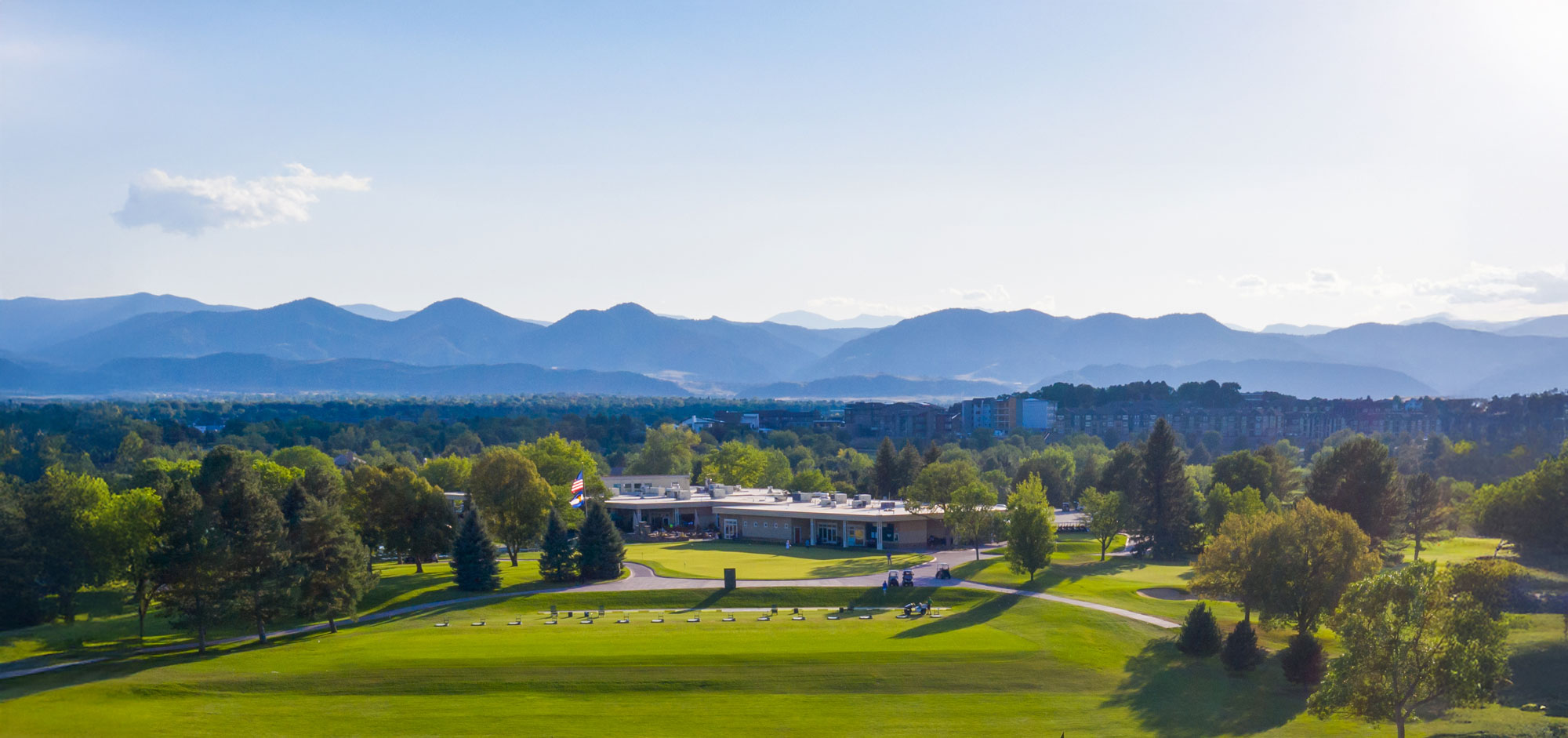 Wide view of a golf course clubhouse surrounded by trees with mountains in the distance under a clear sky.