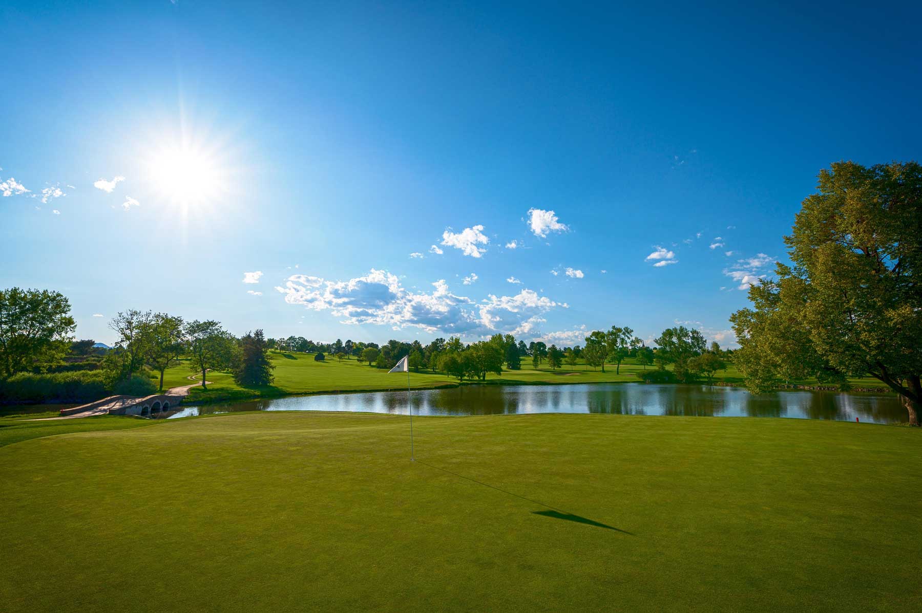 Golf green with flag, reflecting pond, and surrounding trees under a sunny blue sky with scattered clouds.