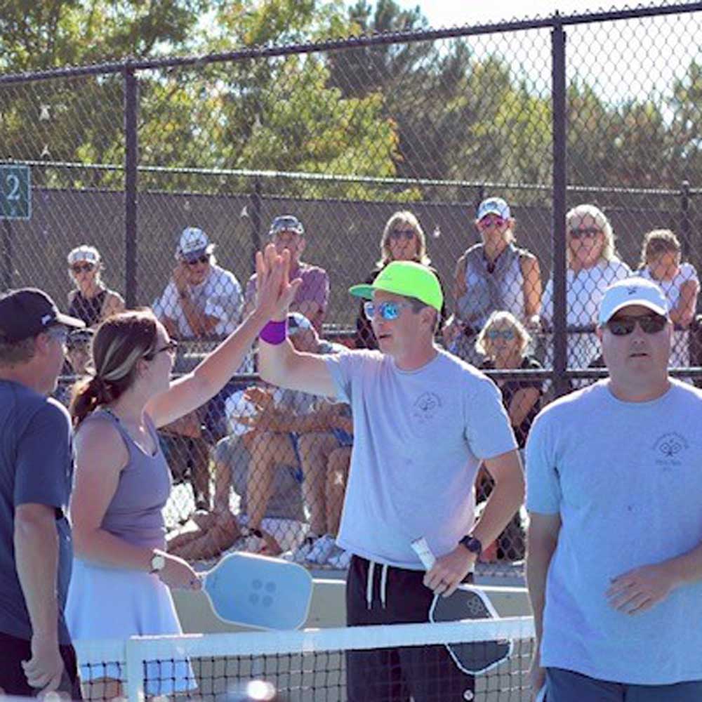Two pickleball players giving a high-five at the net with spectators behind a chain-link fence.