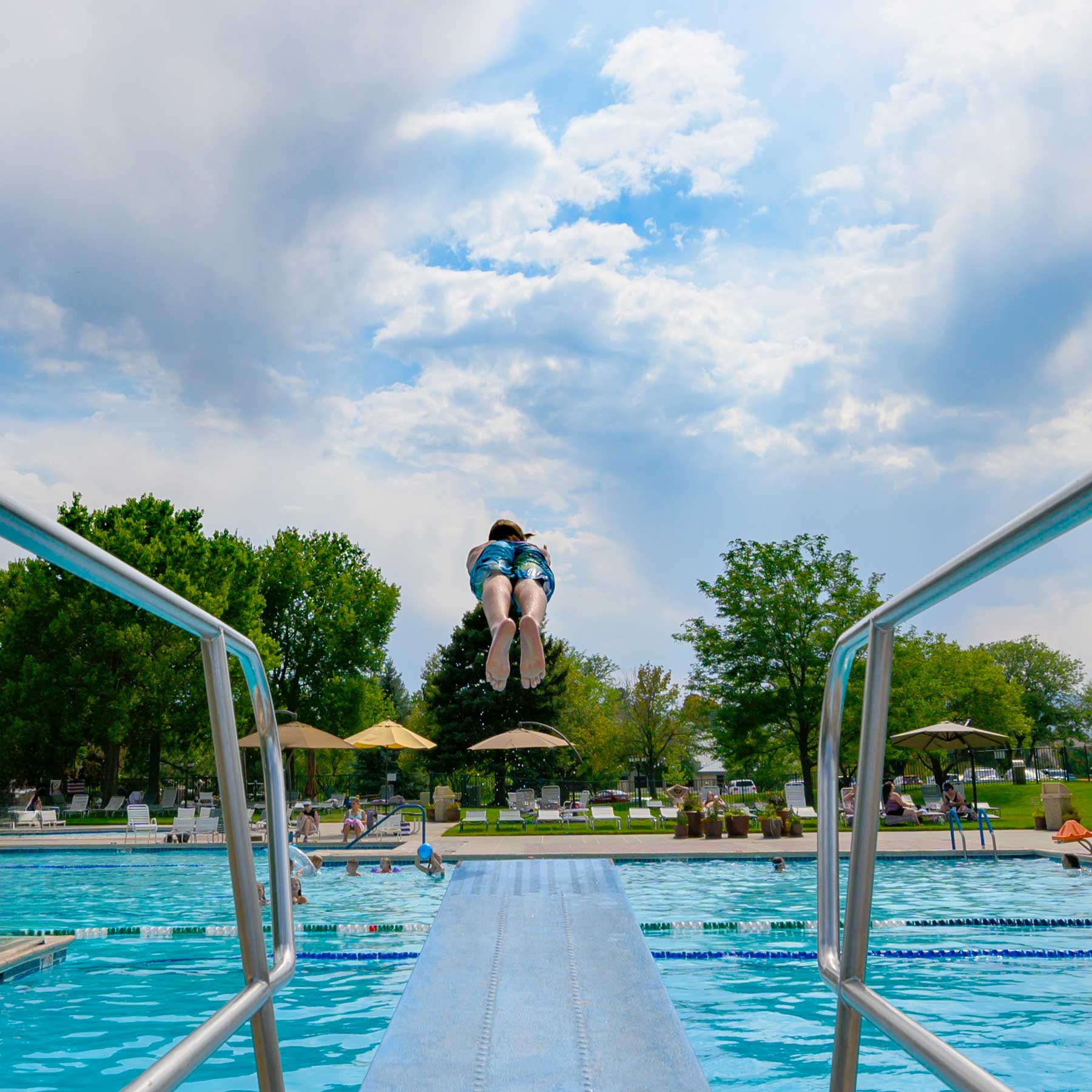 Child in swim trunks mid-air jumping off a diving board into an outdoor pool with lounge chairs, umbrellas, and trees in the background.